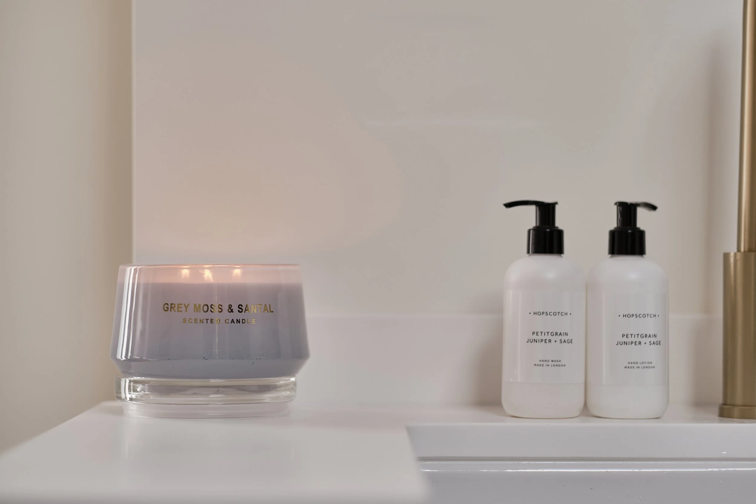A white shelf holding a lit scented candle in a glass container and two white pump bottles labeled hand wash and hand lotion with black pumps, all in a minimalistic bathroom setting.