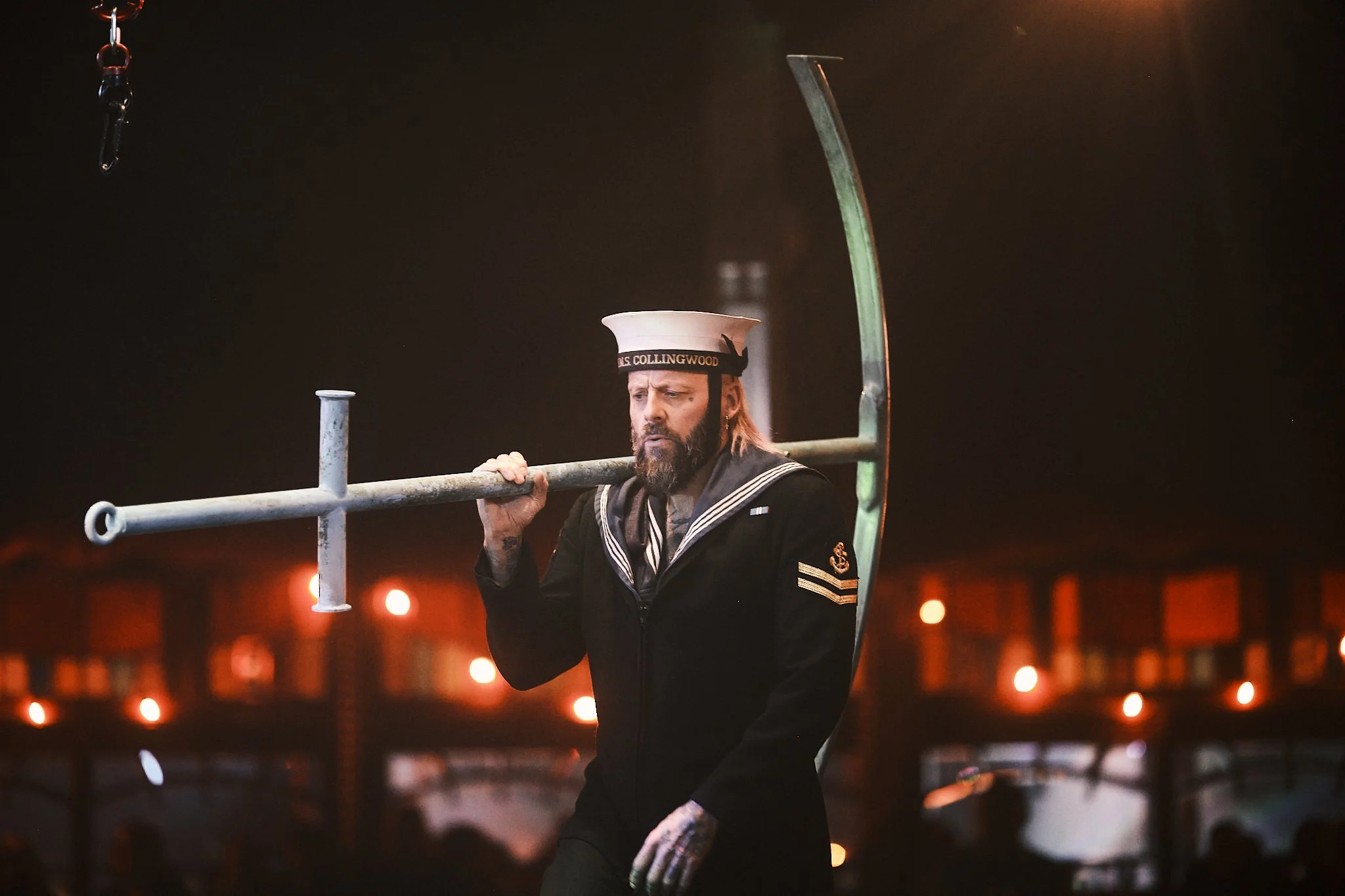 A man dressed in a sailor uniform carrying a metal anchor over his shoulder at night.