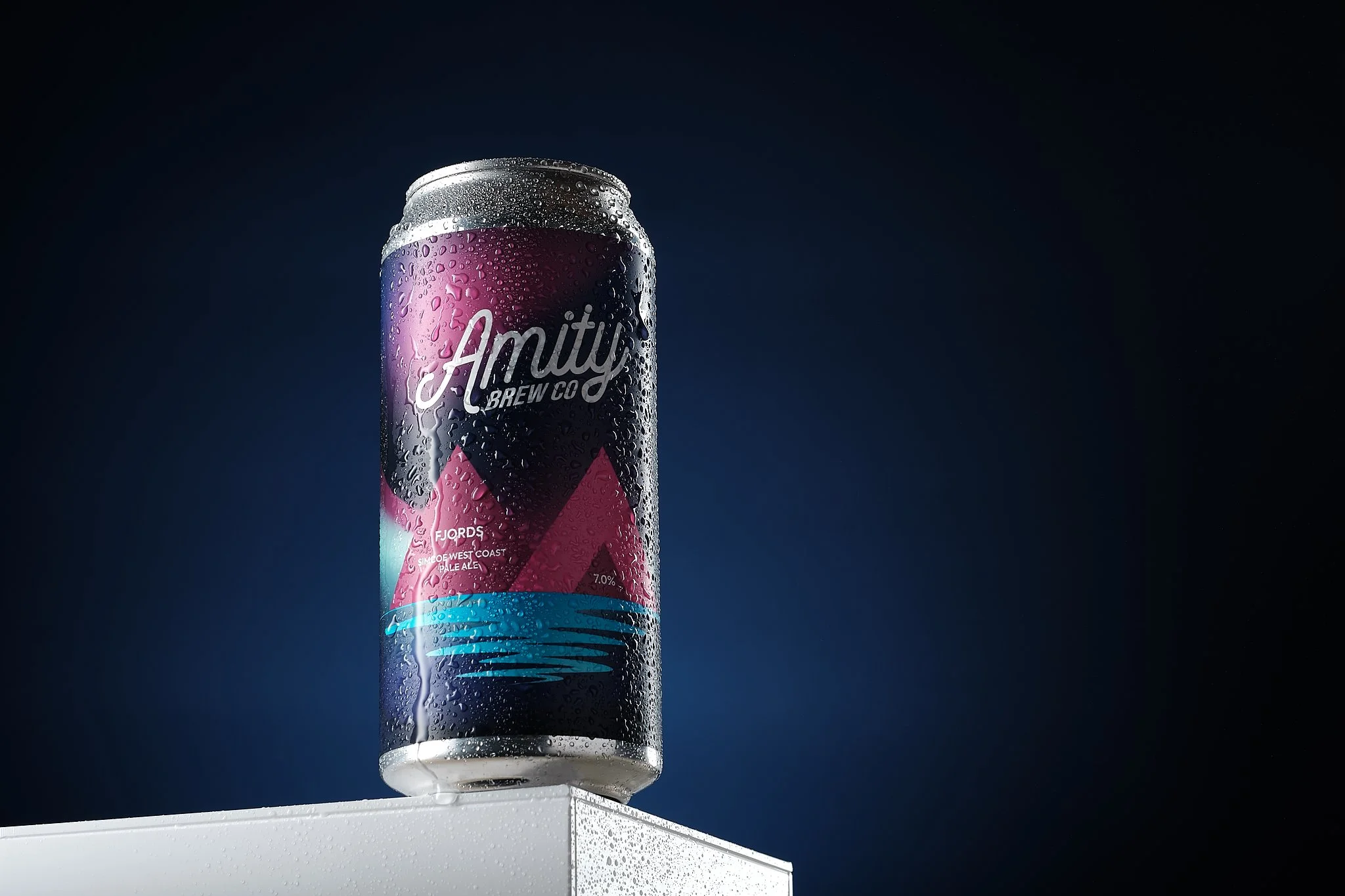 A can of Amity Brew Co. beer rests on a white surface, condensation droplets visible on its surface, against a dark background