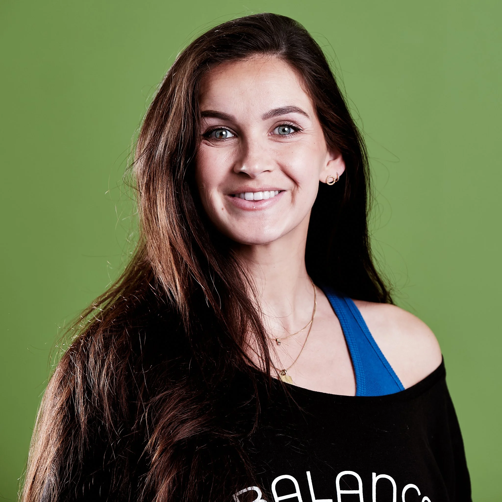 A woman with long dark hair, blue eyes, and jewelry, wearing a black top with a blue strap, smiling against a green background.