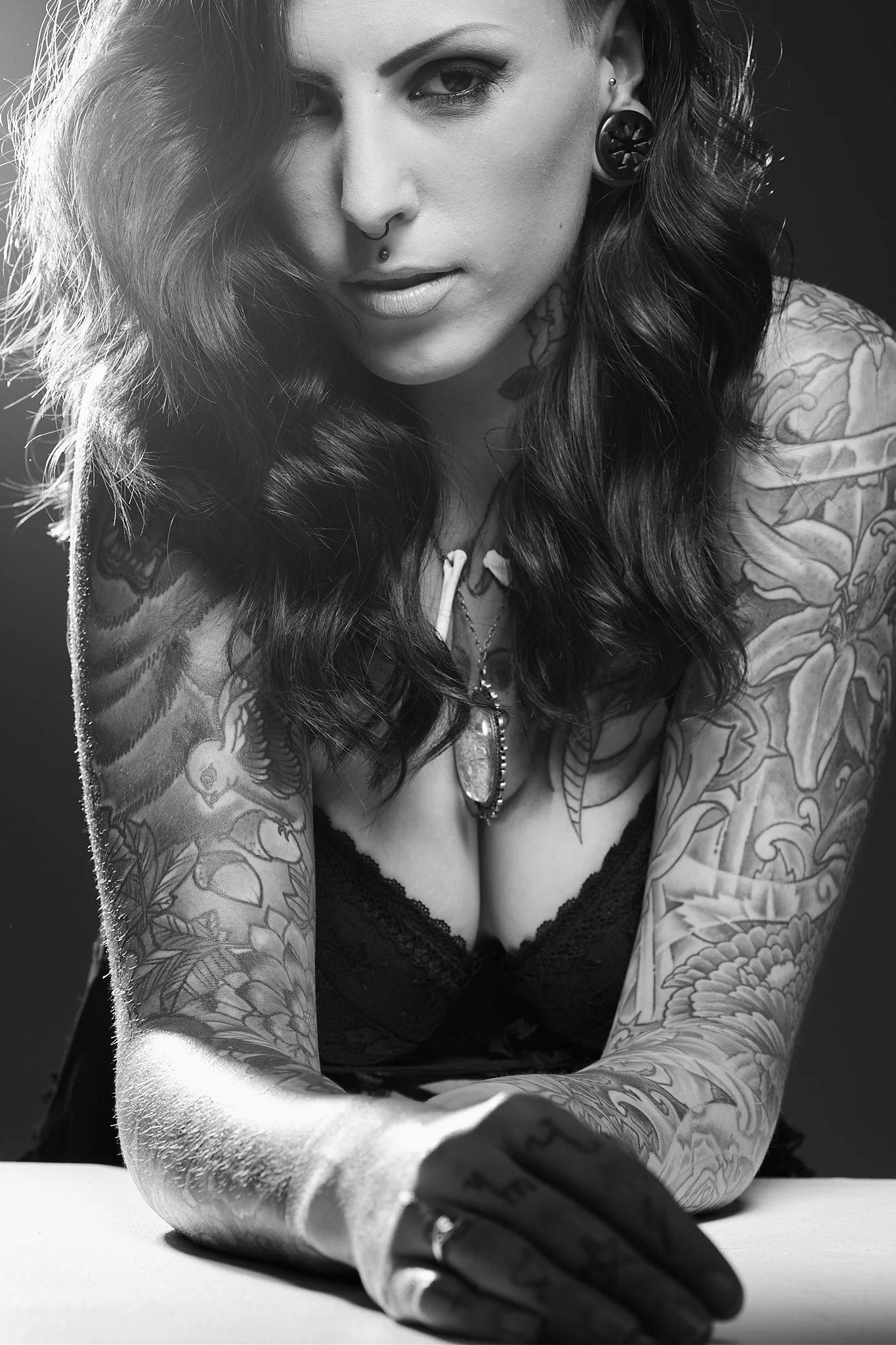 Black and white photo of a tattooed woman with dark wavy hair, wearing a black lace top, leaning forward with her right arm resting on a surface, showing tattoos on her arm, shoulder, and chest, and wearing earrings and a nose ring.