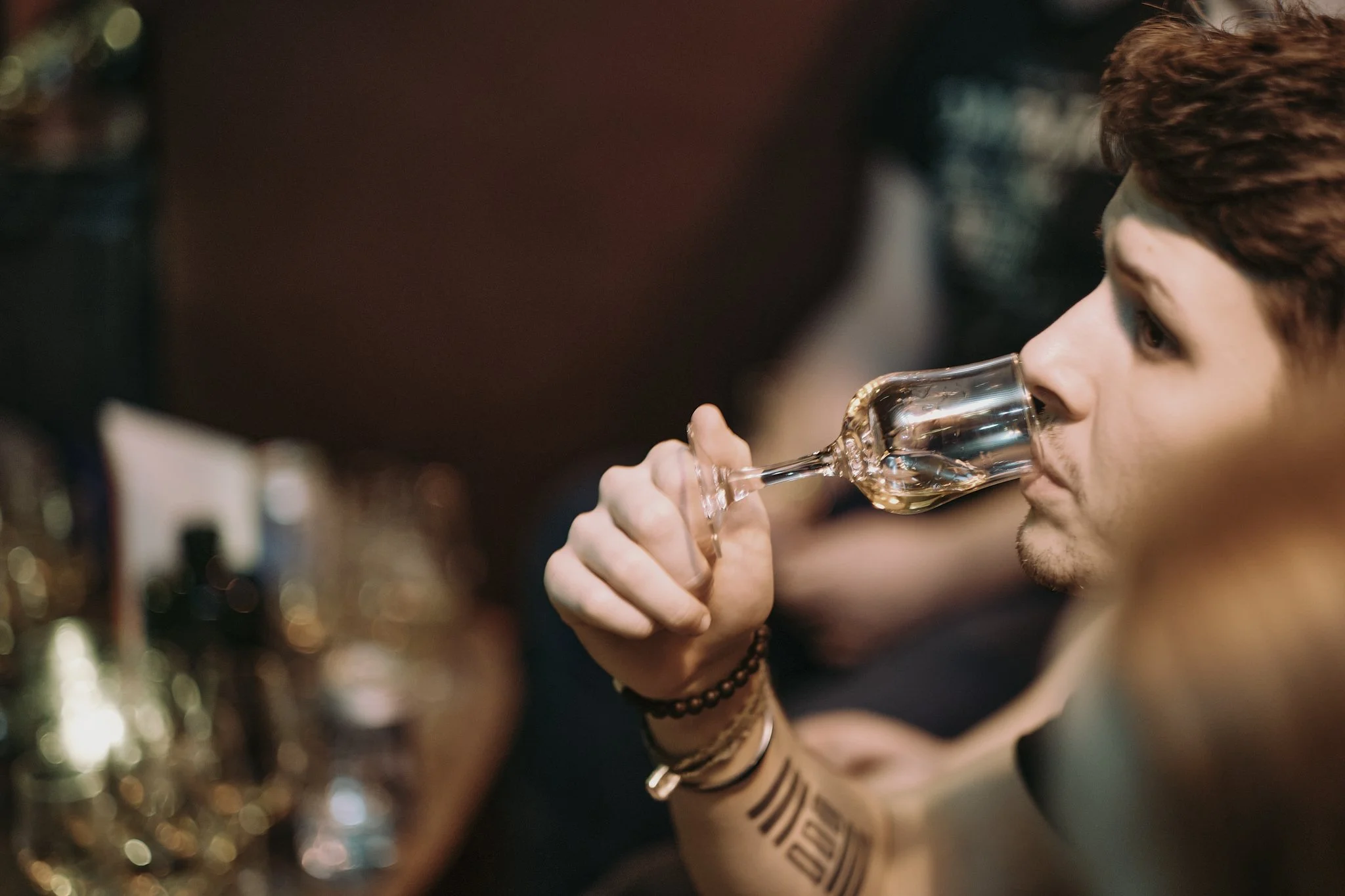 A man with short dark hair and tattoos on his forearm sipping from a clear glass wine glass at a bar or social setting.