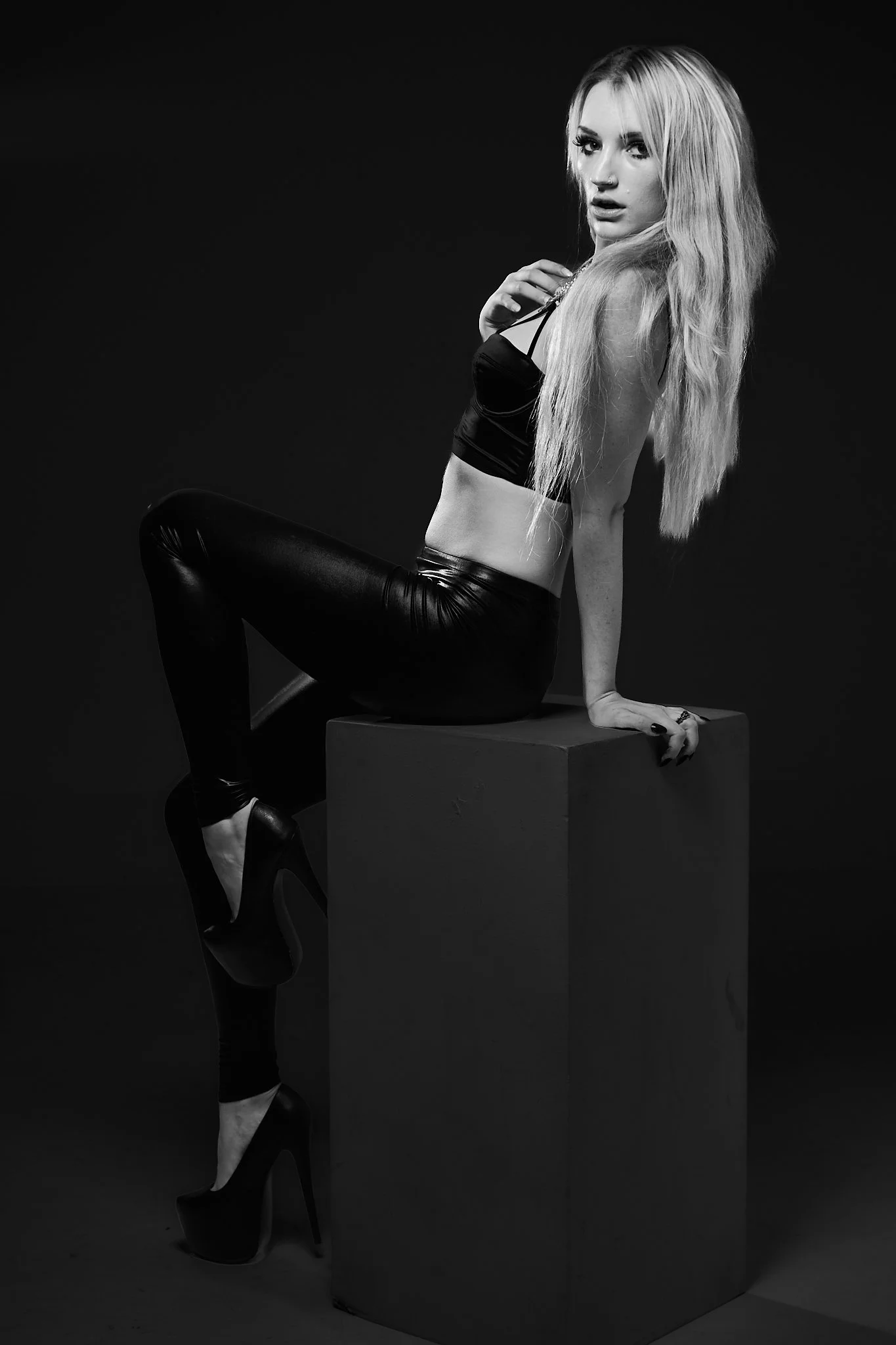 A black and white photo of a woman with long, light-colored hair sitting on a rectangular block, wearing a crop top, leather pants, and high heels.