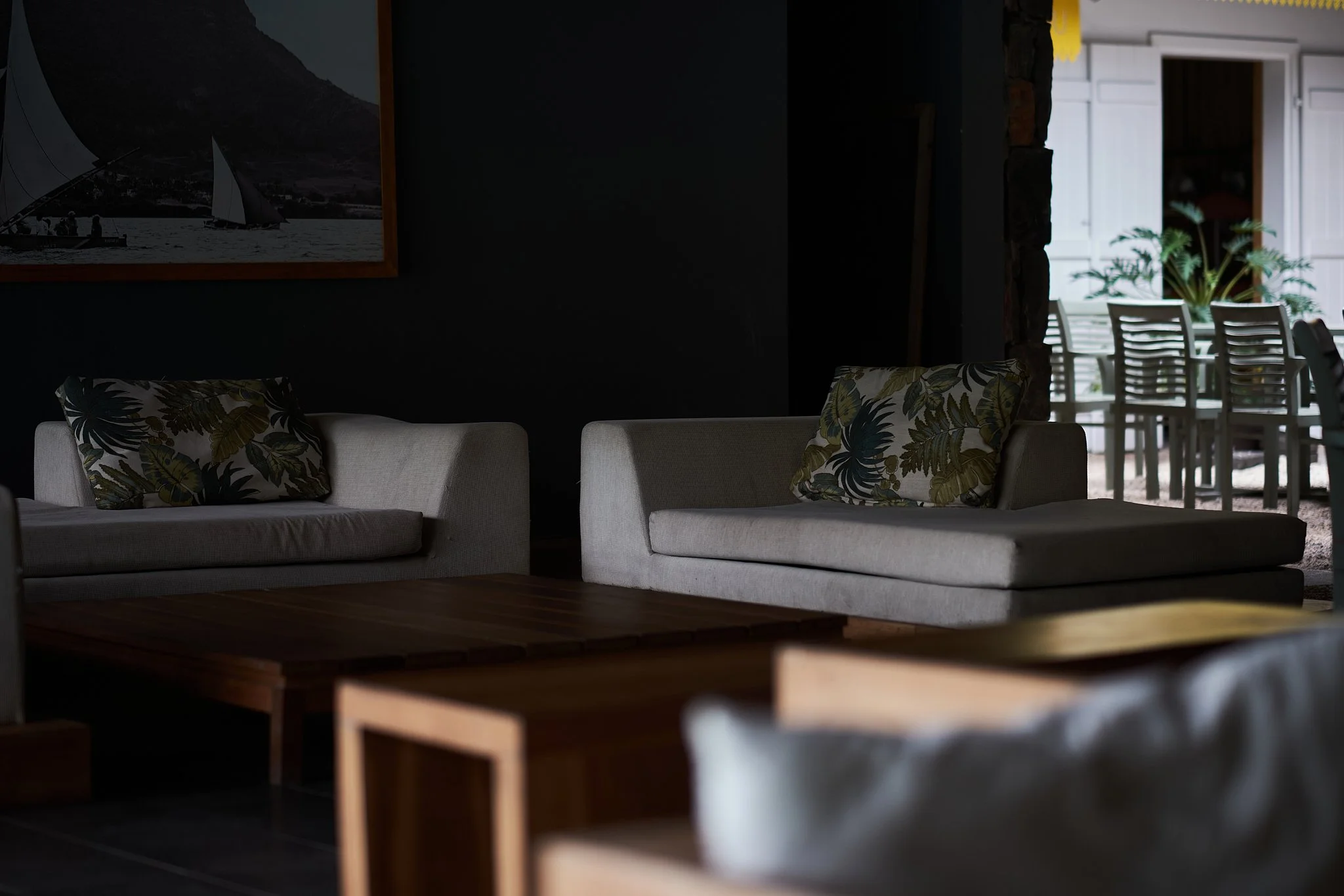 Living room with two light-colored sofas, each with tropical-patterned pillows, a wooden coffee table, and a dark wall with a framed boat or sailboat picture.