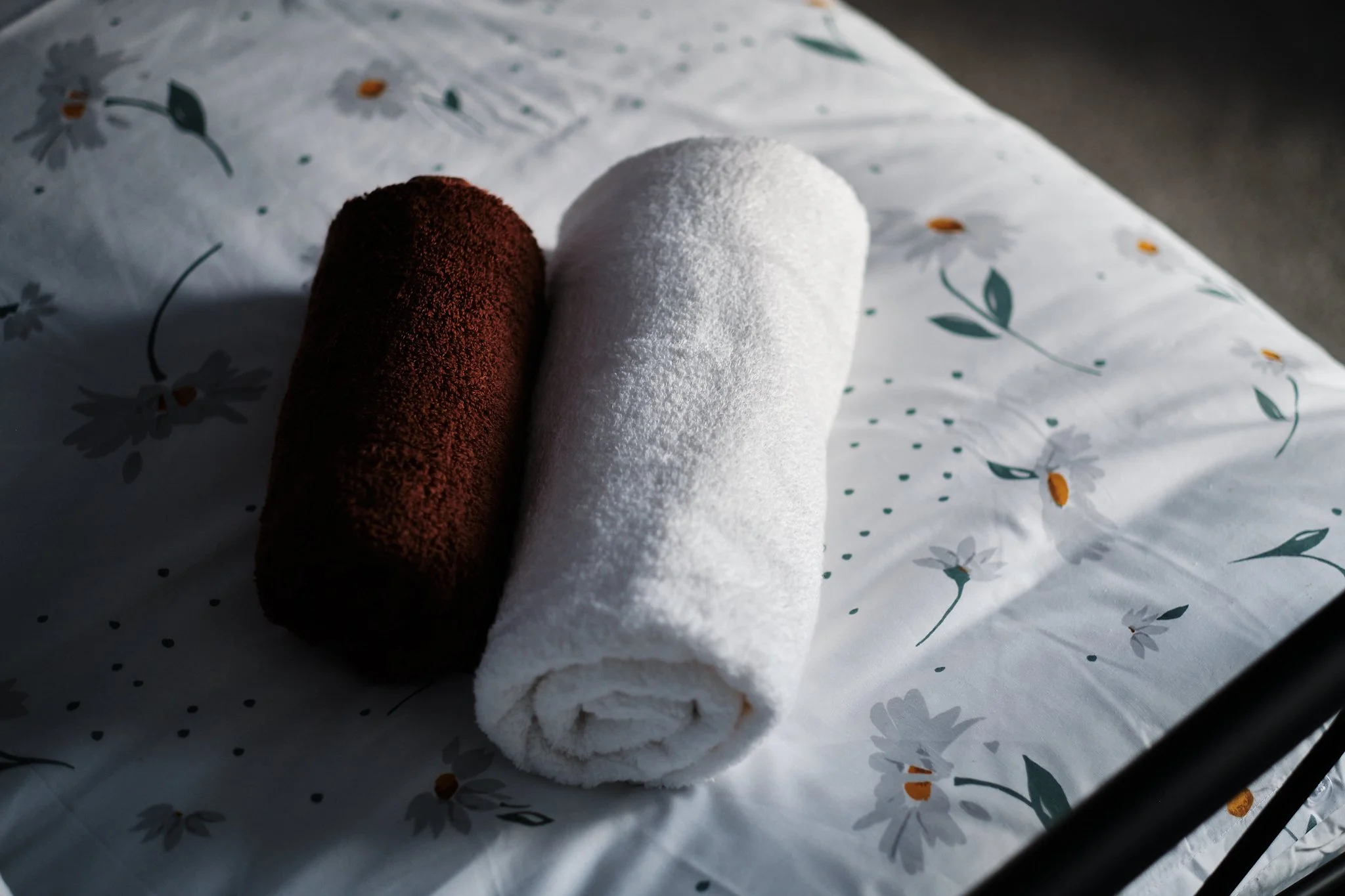 A brown towel and a white towel rolled up, placed on a floral patterned bed sheet.