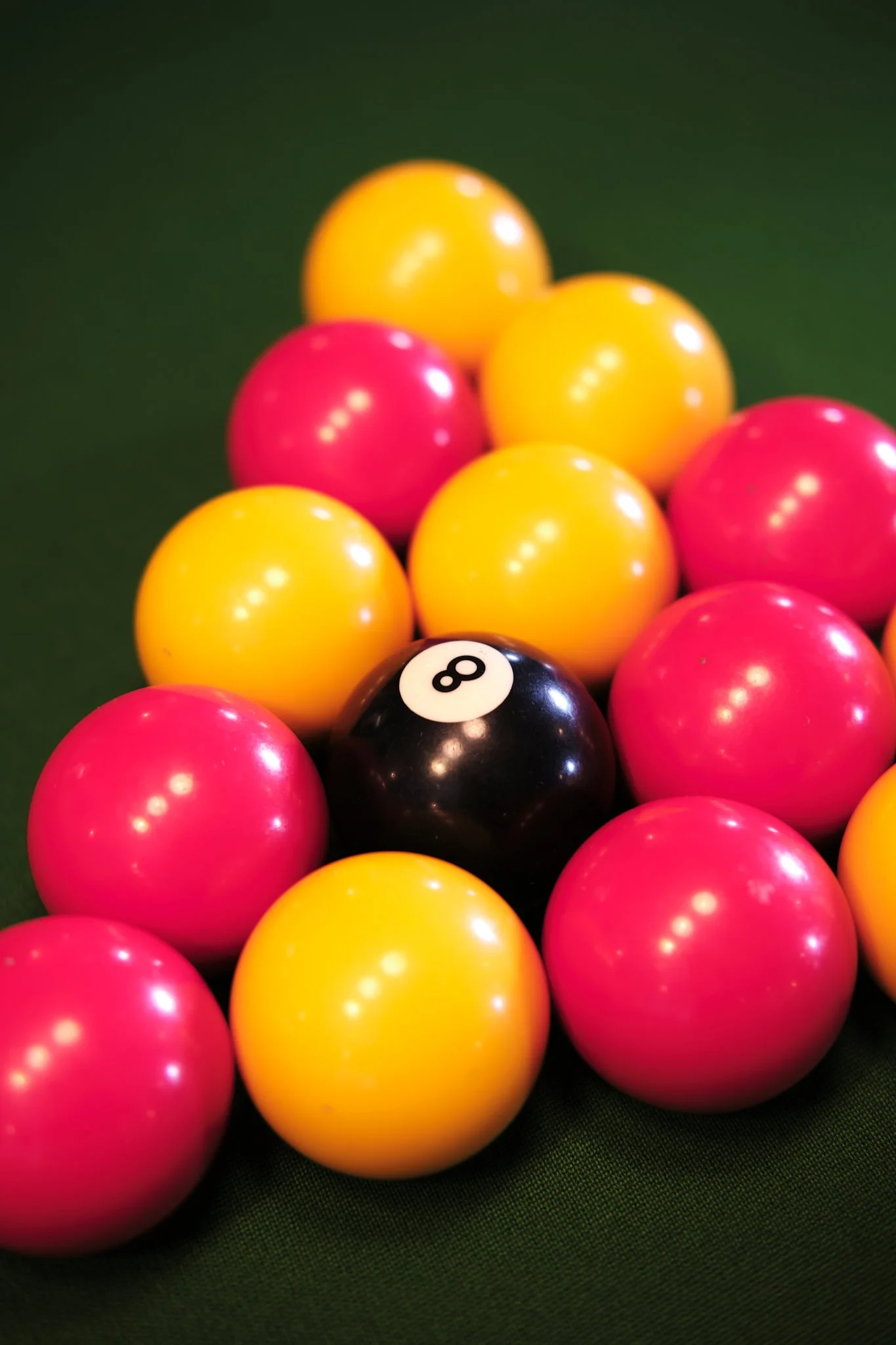 A set of pink, yellow, and black billiard balls arranged on a green pool table, with the 8 ball in the center.