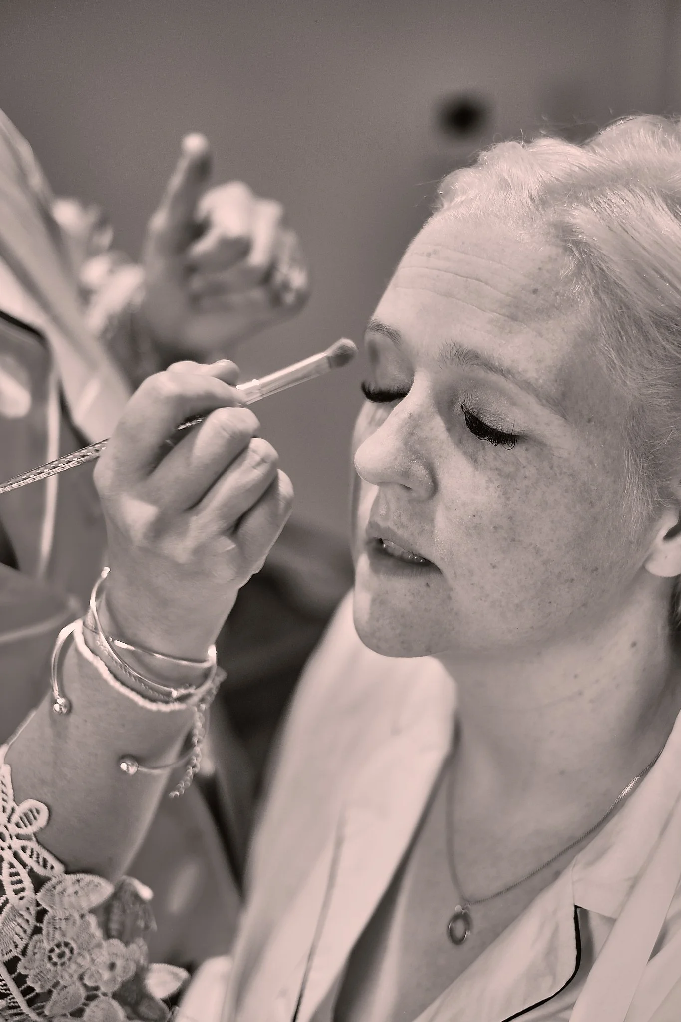 A woman with light hair and closed eyes wearing a dress with lace detail is having her makeup applied with a brush by a makeup artist.