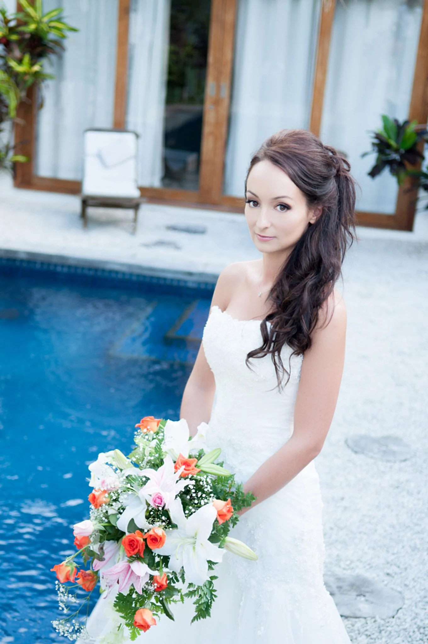 A woman in a white wedding dress standing outdoors near a swimming pool, holding a large bouquet of colorful flowers, with a wooden building and glass doors in the background.
