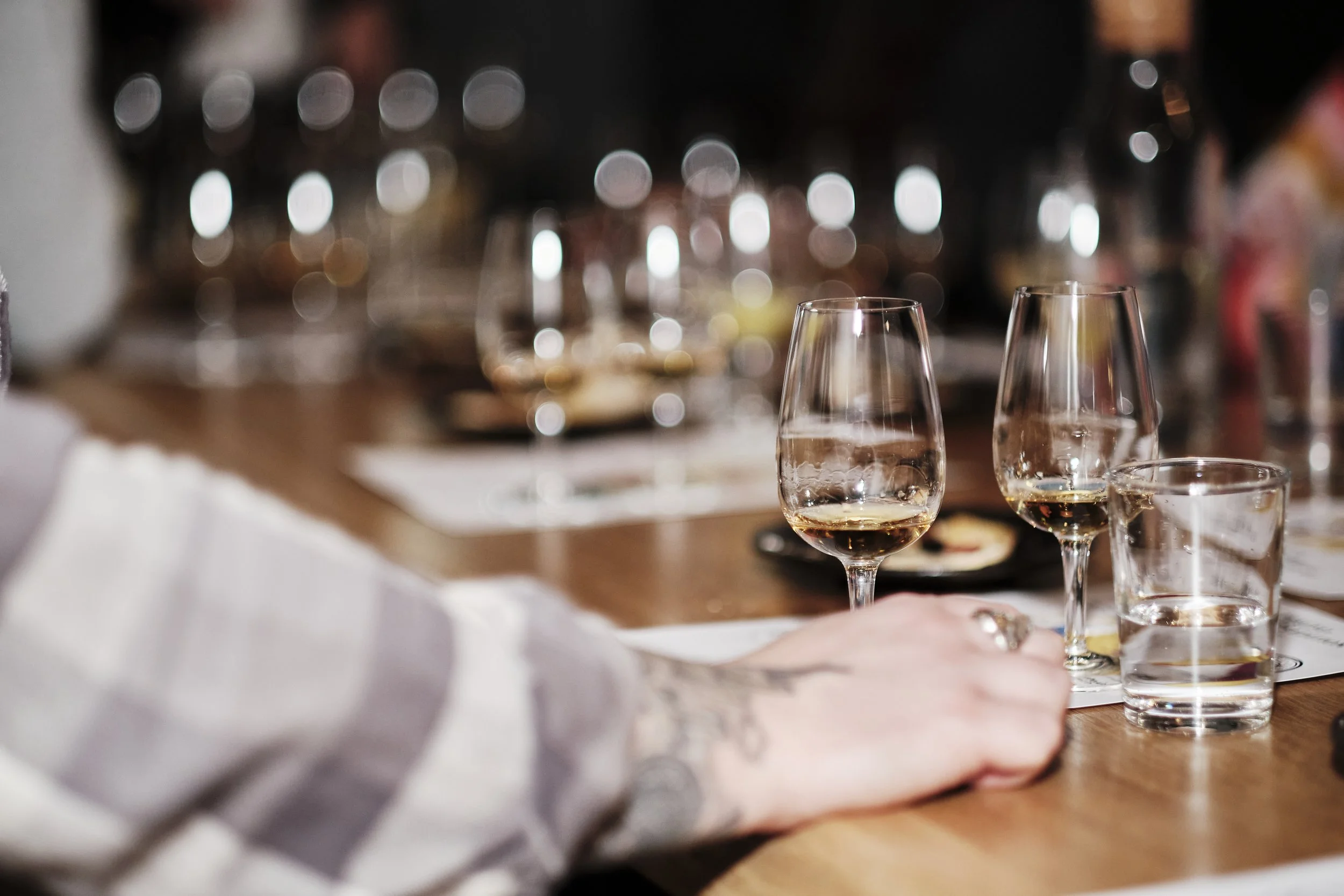 Close-up of a hand resting on a table with several wine glasses and a glass of water in front, blurred background with more glasses and a buffet or dining setup.