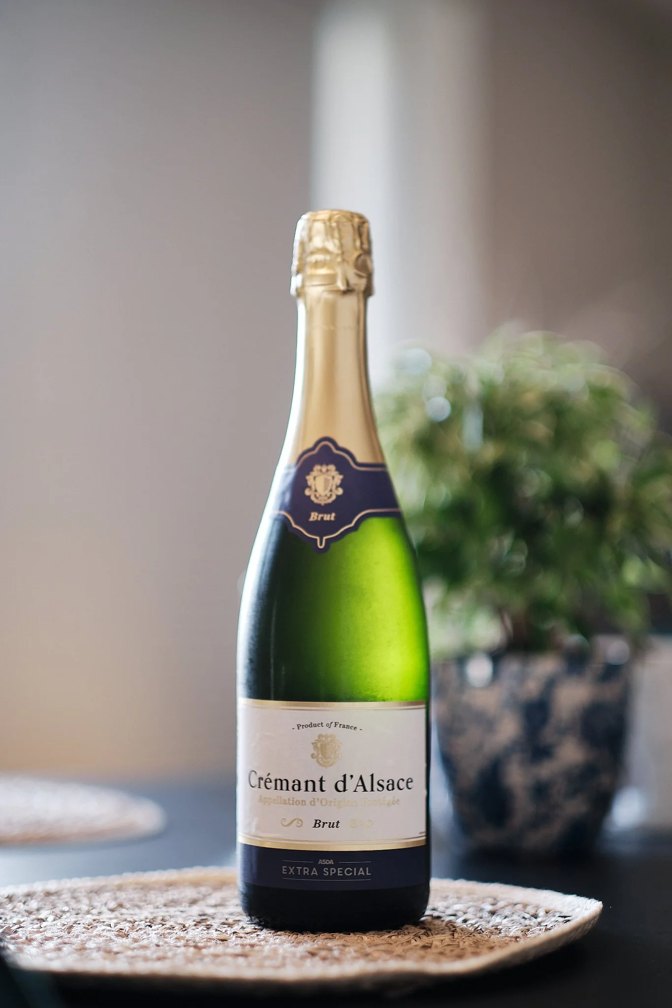 A bottle of Crémant d'Alsace sparkling wine on a woven placemat, with a blurred potted plant and neutral background.