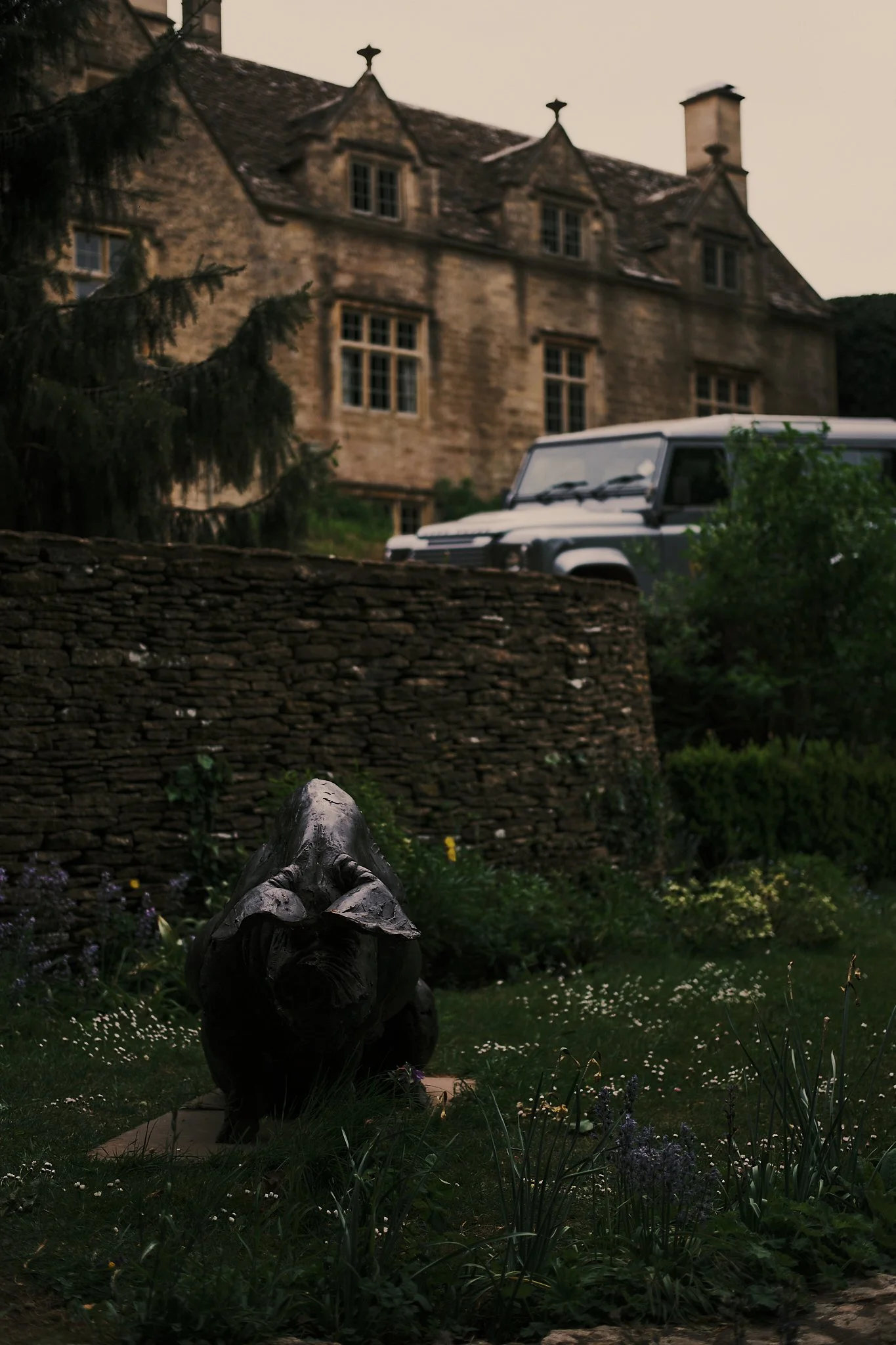 A garden with a pig sculpture, greenery, and flowers in front of a stone wall, with an old house and a land rover in the background. Hotel interior photography at THE PIG-in the Cotswolds
