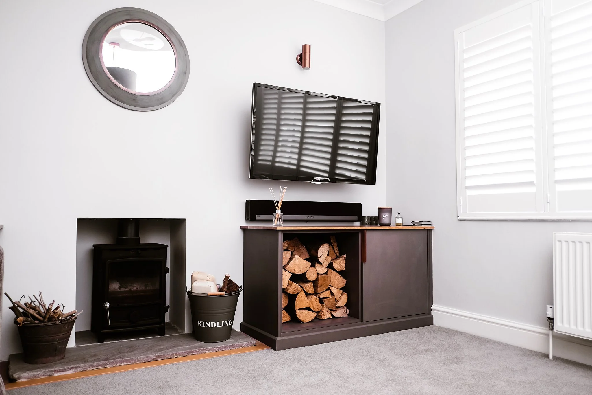 A living room corner with a white wall, a mounted flat-screen TV, a window with white shutters, a black fireplace, a basket of firewood and kindling, a ceiling light, and a dark wood cabinet with stacked firewood, a soundbar, candles, and decor items