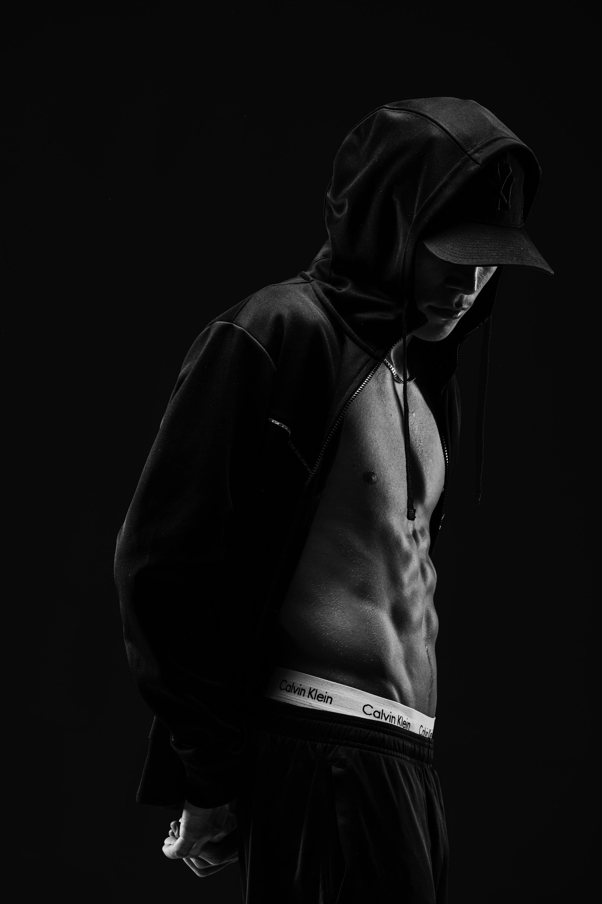 Black and white photo of a shirtless man wearing a hoodie, cap, and Calvin Klein underwear, with his head down, against a dark background.