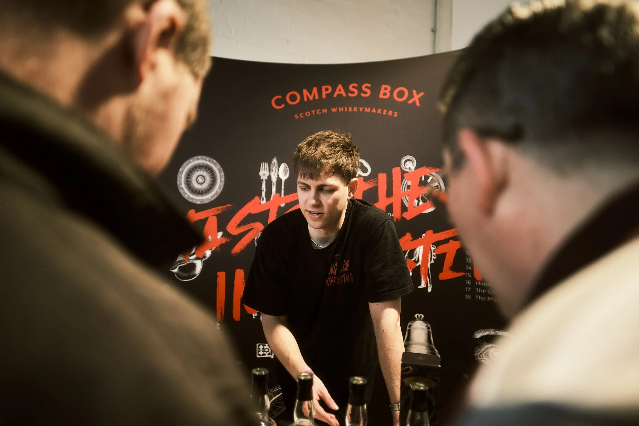 A young man demonstrating whisky tasting at a whisky event with a black backdrop reading 'Compass Box Scotch Whisky Makers' and 'Taste the Untasted' in red text, surrounded by a few onlookers.