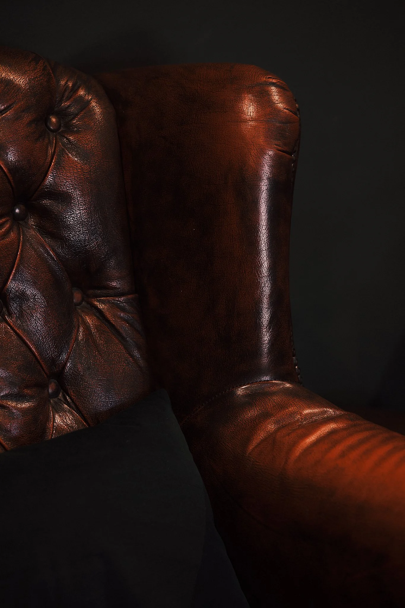 Close-up of a brown leather tufted armchair with a black pillow nearby, set against a dark background.