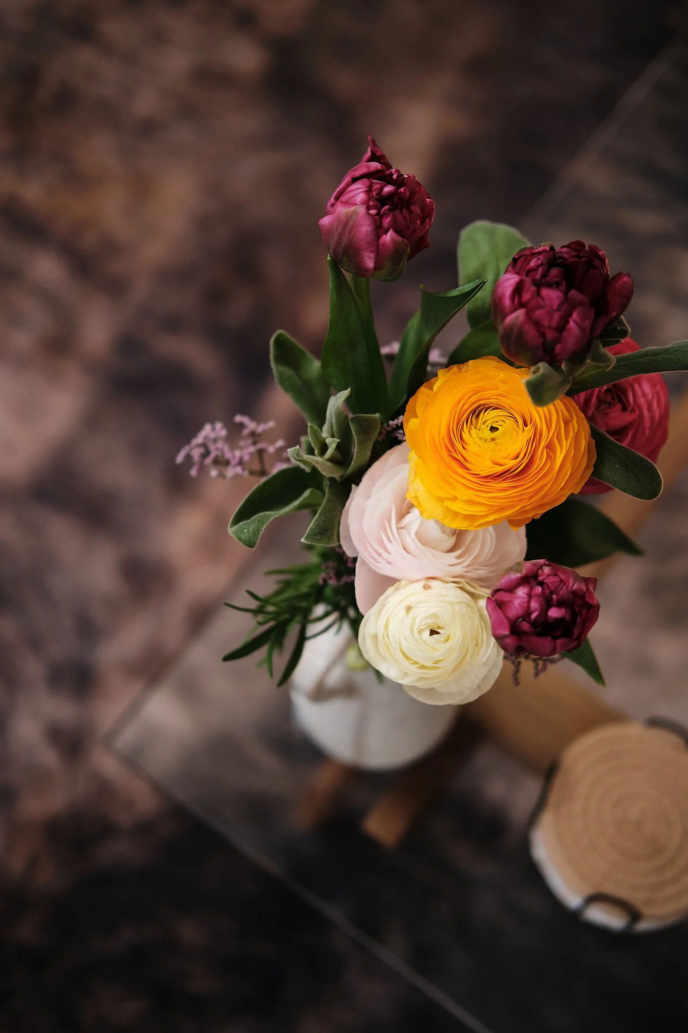A bouquet of assorted flowers in a white vase on a glass table with a wooden piece nearby.