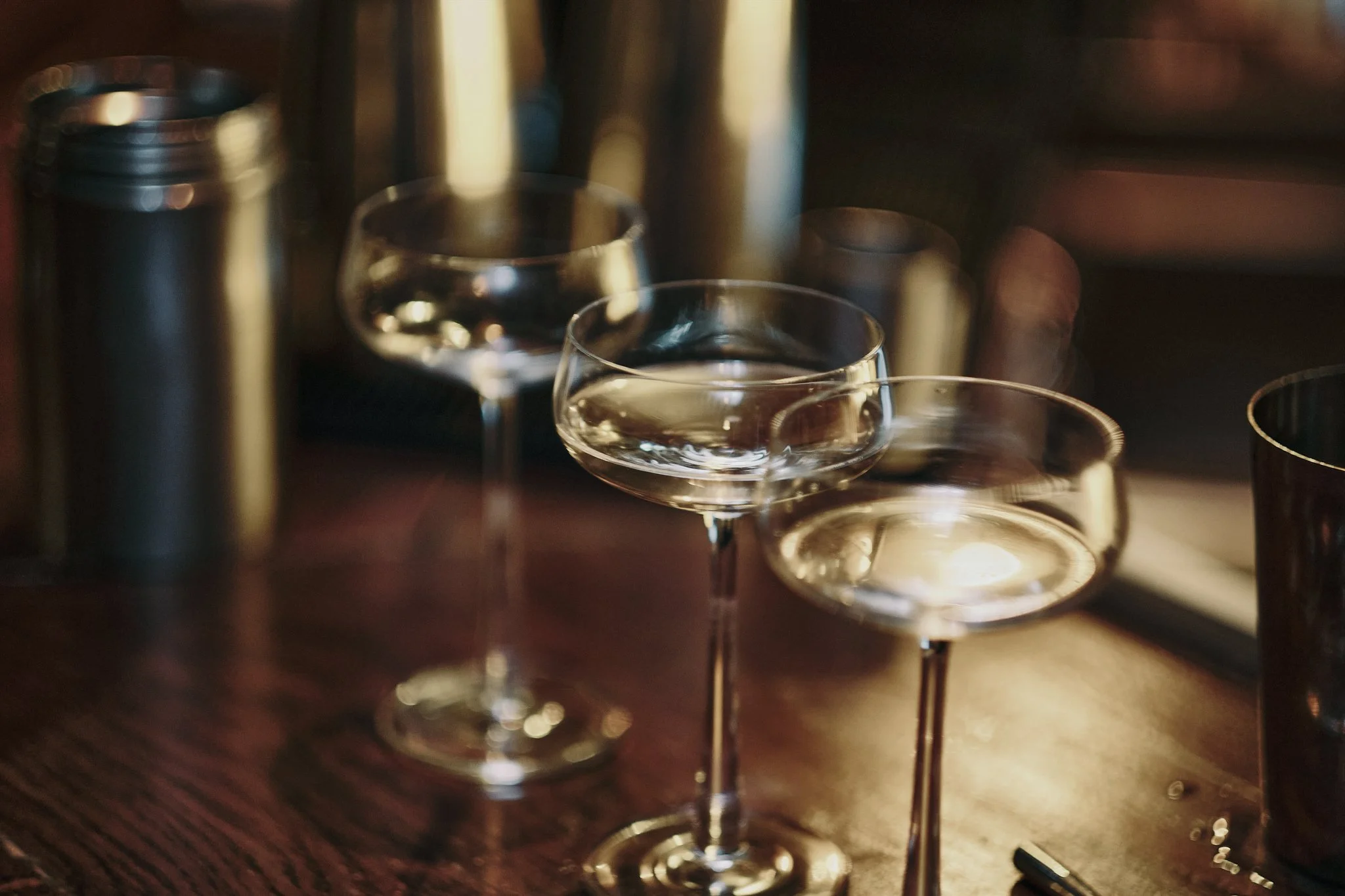 Three coupe glasses with clear liquid on a wooden table, with blurred bottles and glasses in the background.