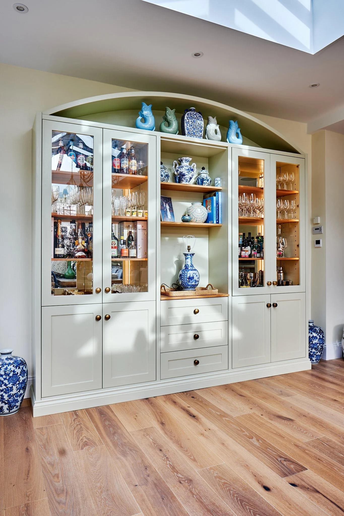 A white cabinet with glass doors displaying various bottles and glassware, topped with blue and white porcelain vases and decorative items, in a room with wooden flooring and a skylight overhead.