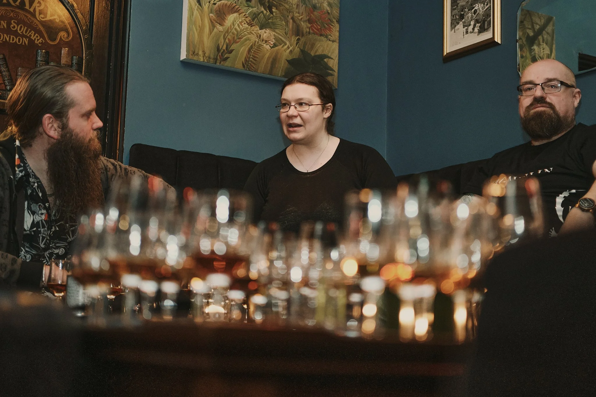 Three people sitting on a dark-colored couch in a room with blue walls, engaged in a conversation. There are multiple glasses of drinks on a tray in the foreground.
