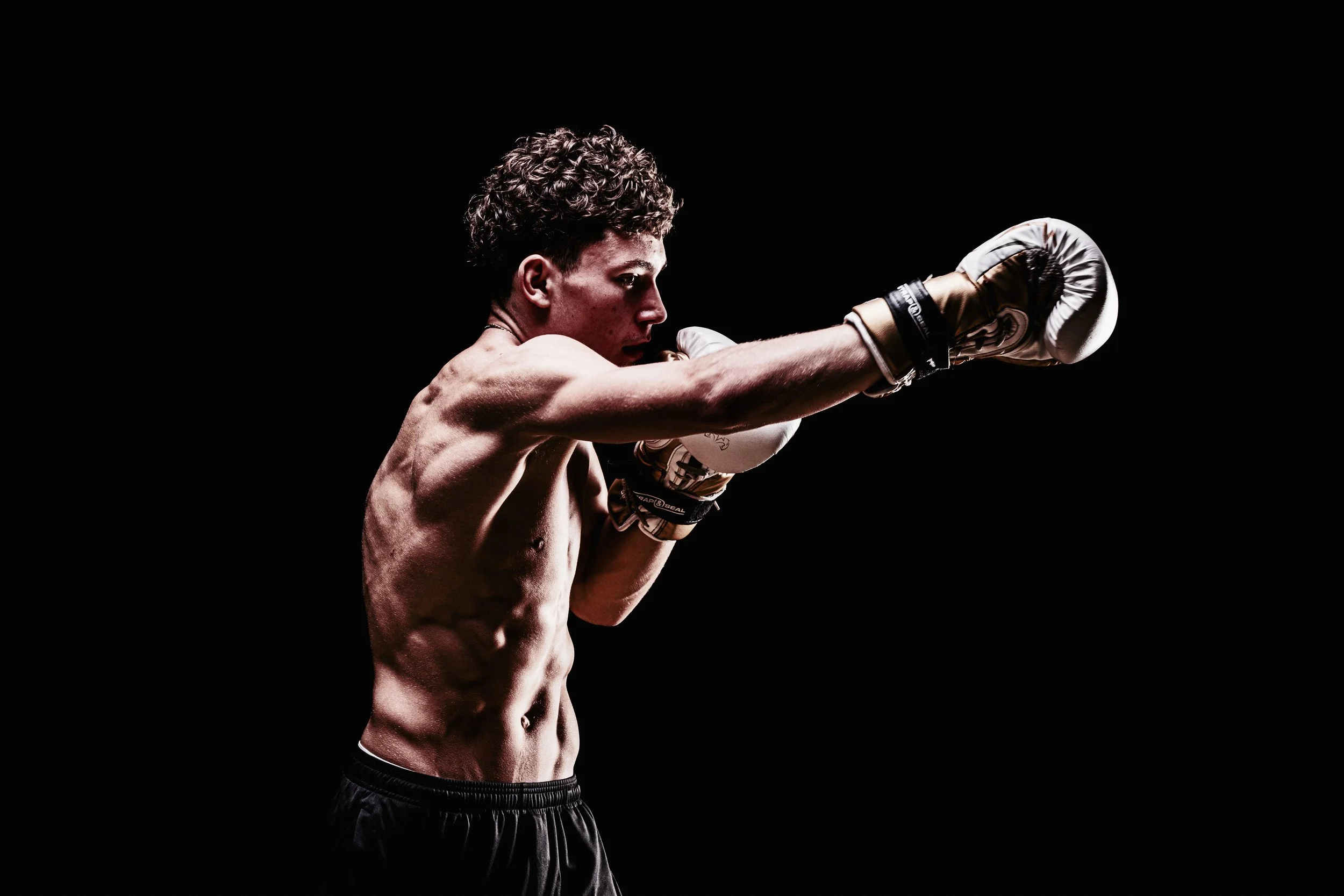 A shirtless male boxer in a fighting stance, wearing boxing gloves, against a black background.