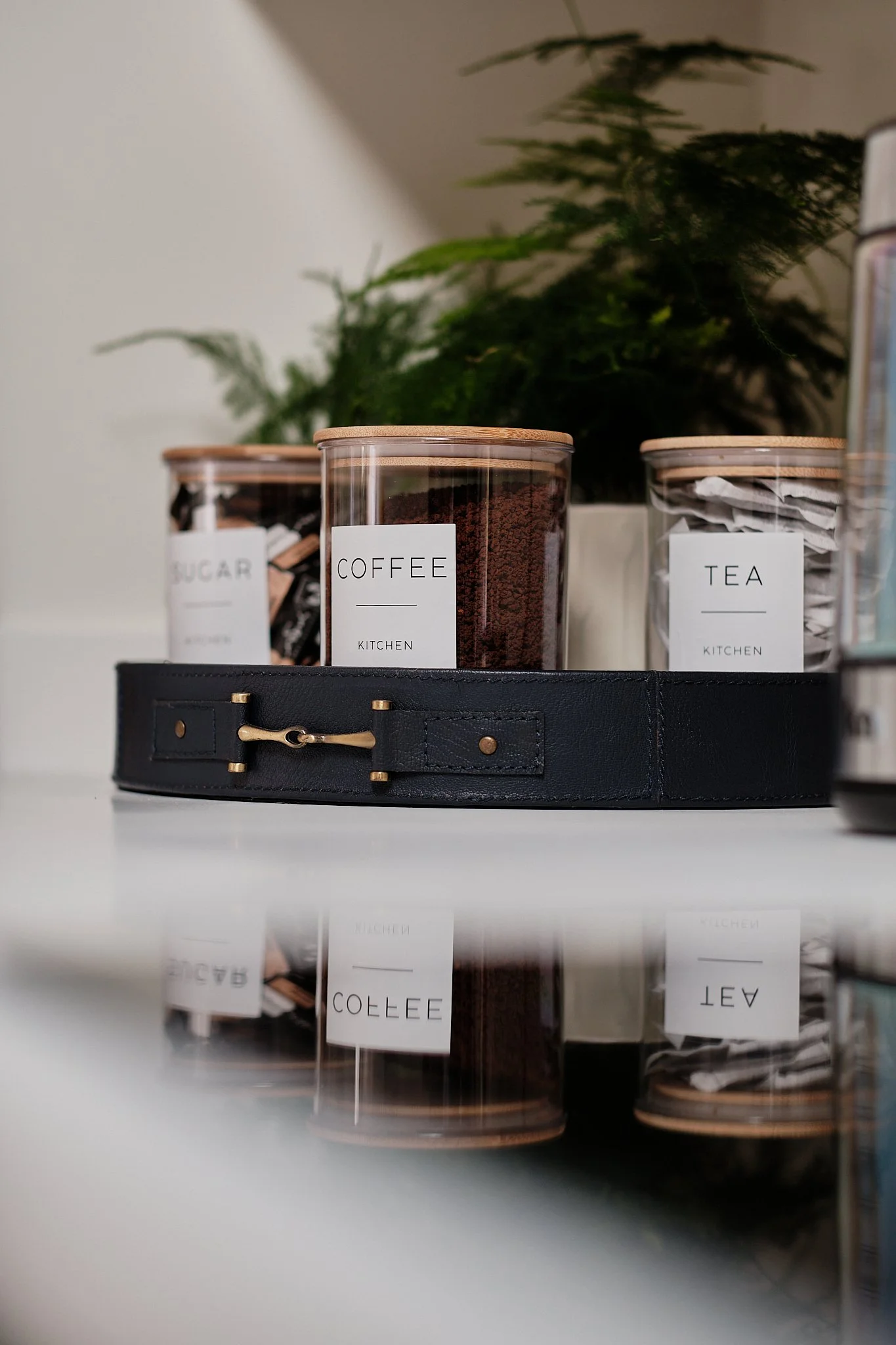 Containers of sugar, coffee, and tea on a reflective surface with a black belt and green plant in the background.