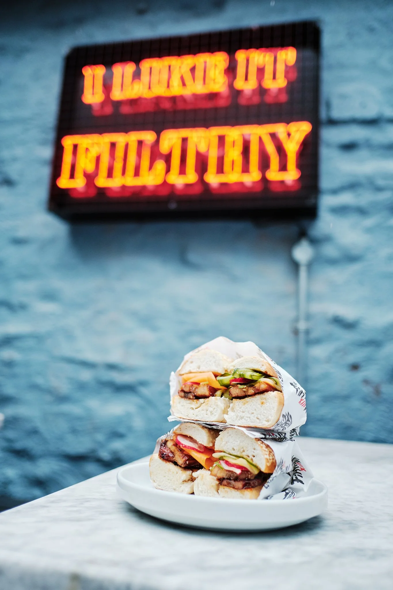 Two sandwiches on a white plate with a brick wall and neon sign that reads 'I Love It' and 'Policy' in the background.