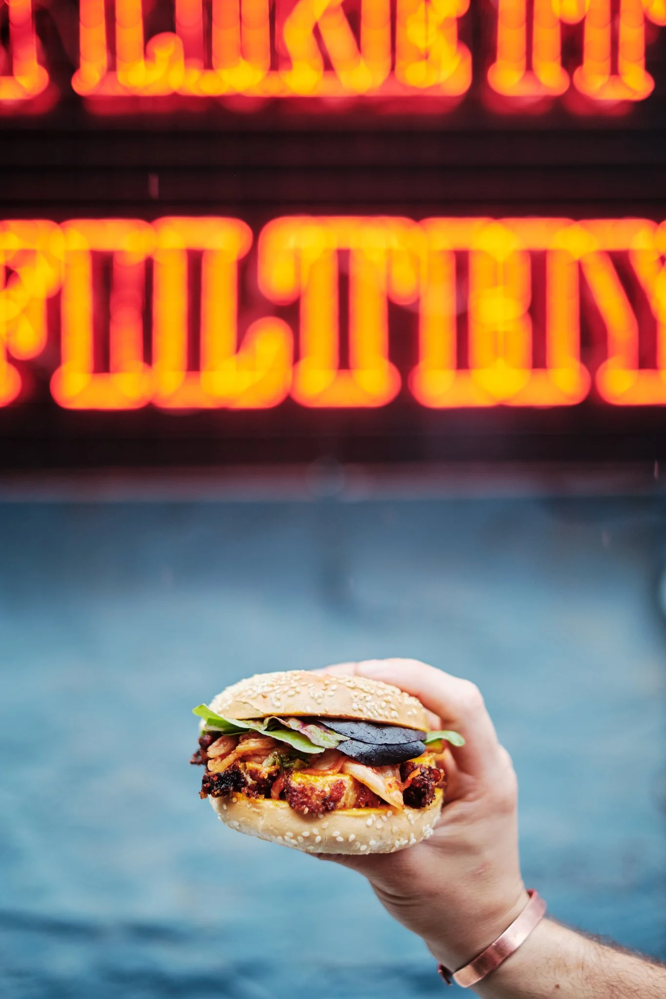 Person holding a burger with lettuce, bacon, and toppings in front of a brightly lit Neon sign.