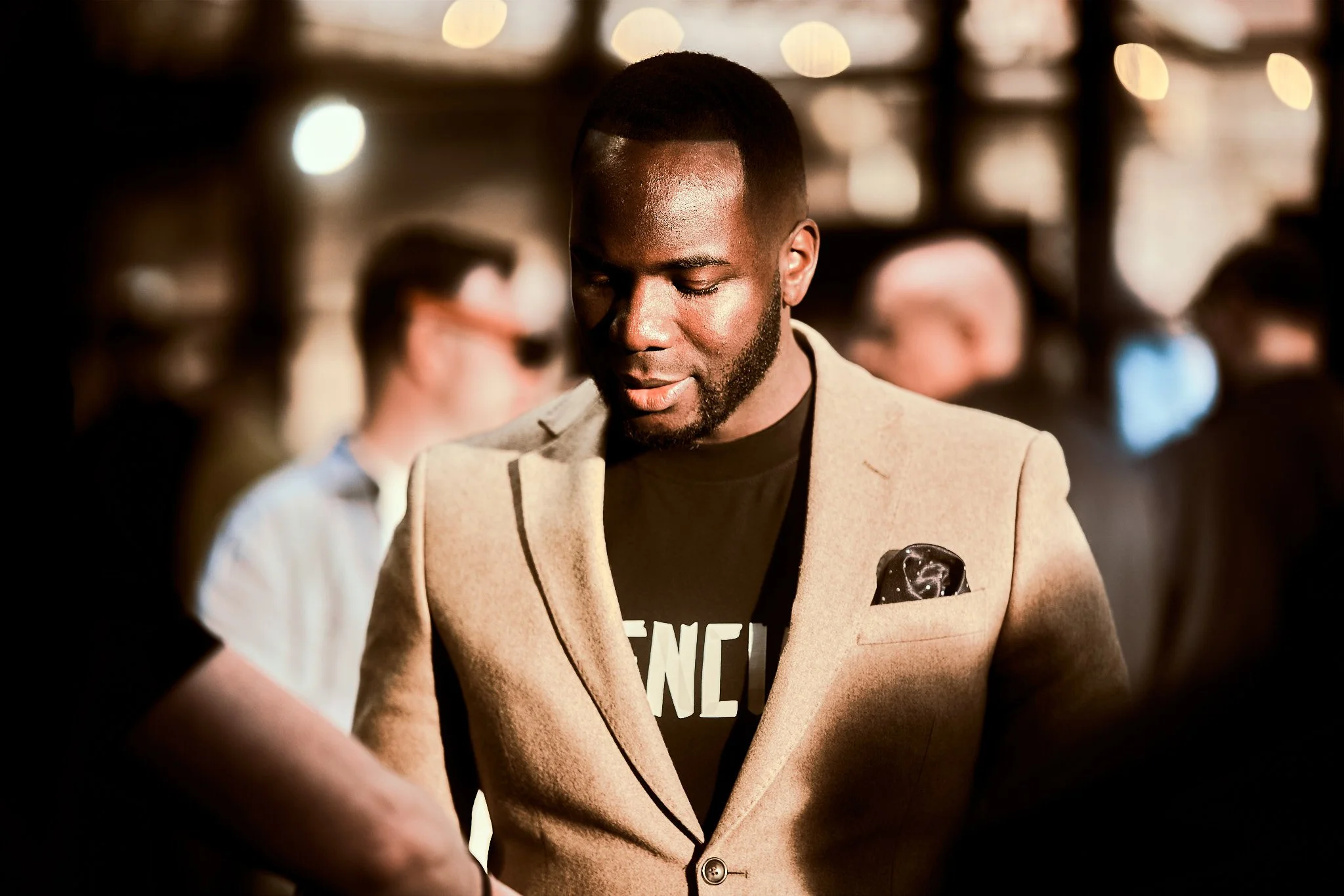 A man wearing a beige blazer with a pocket square, black T-shirt, and earrings, looking down in a dimly lit social setting with blurred background of people.