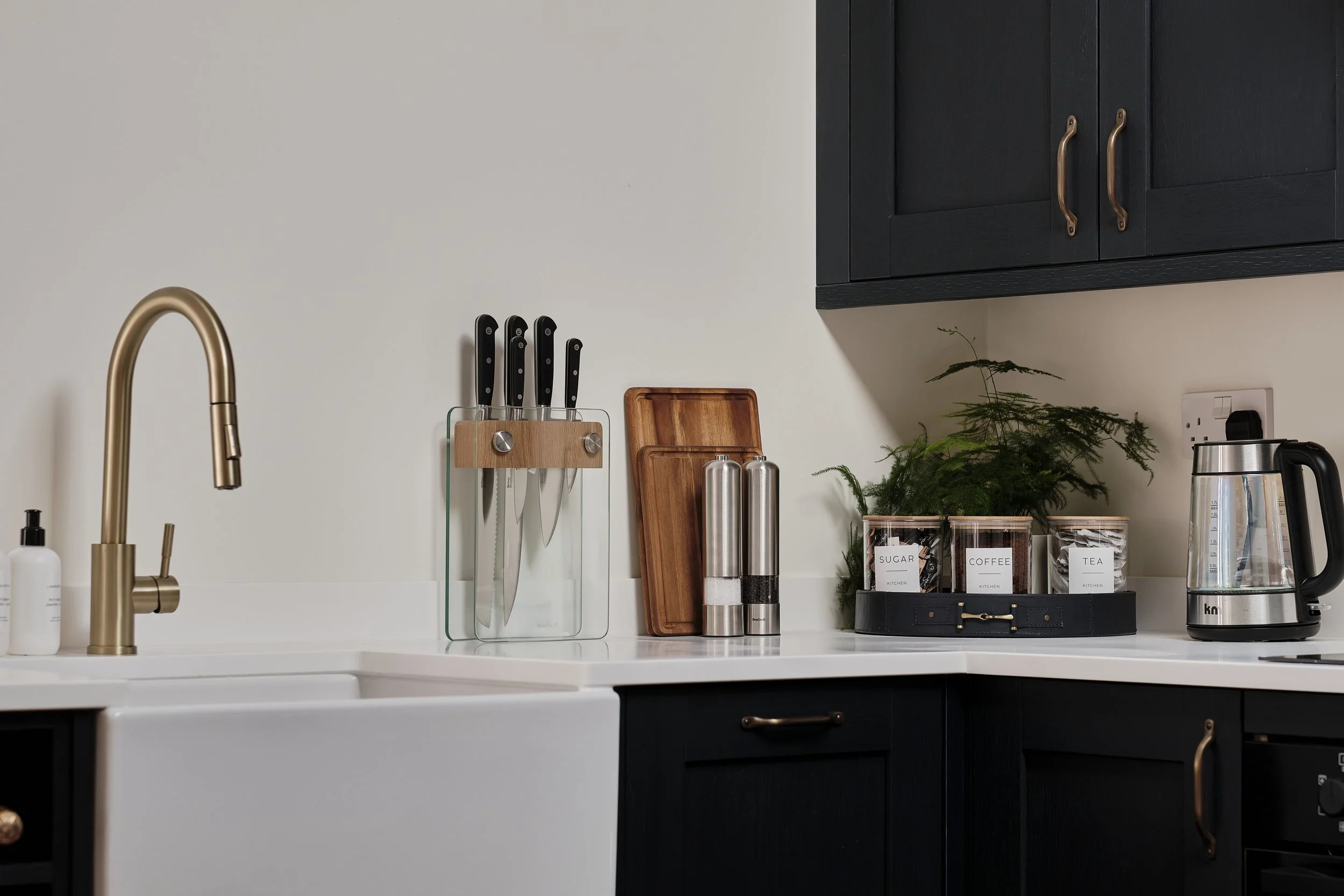 Kitchen countertop with a gold faucet, a white sink, a knife block with black-handled knives, wooden cutting boards, stainless steel salt and pepper shakers, a plant, labeled jars for sugar, coffee, and tea, and a glass electric kettle, with dark cab