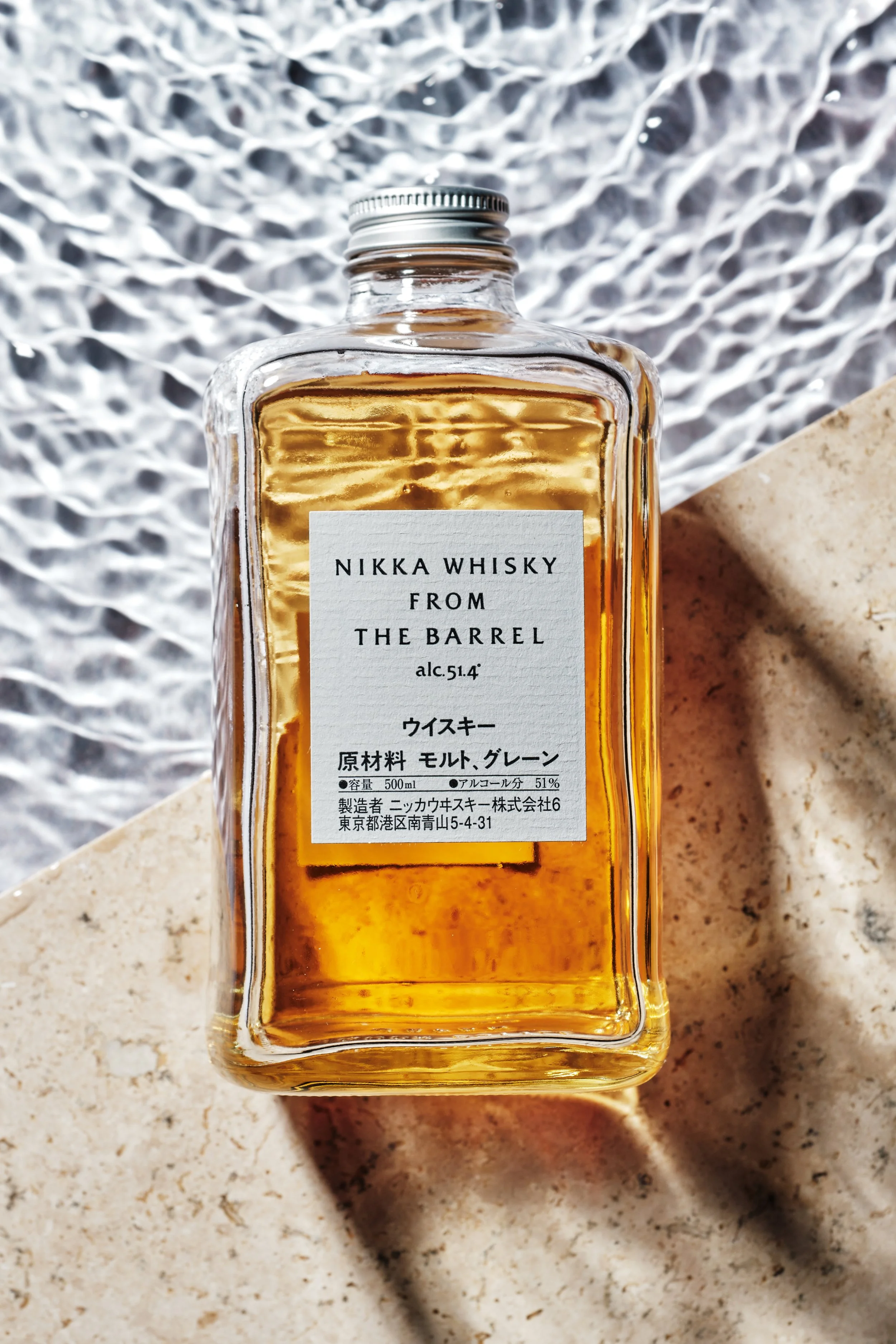 A bottle of Nikka Whisky from the Barrel, with a silver cap, filled with amber-colored whisky, placed on a stone surface with a textured metal background.