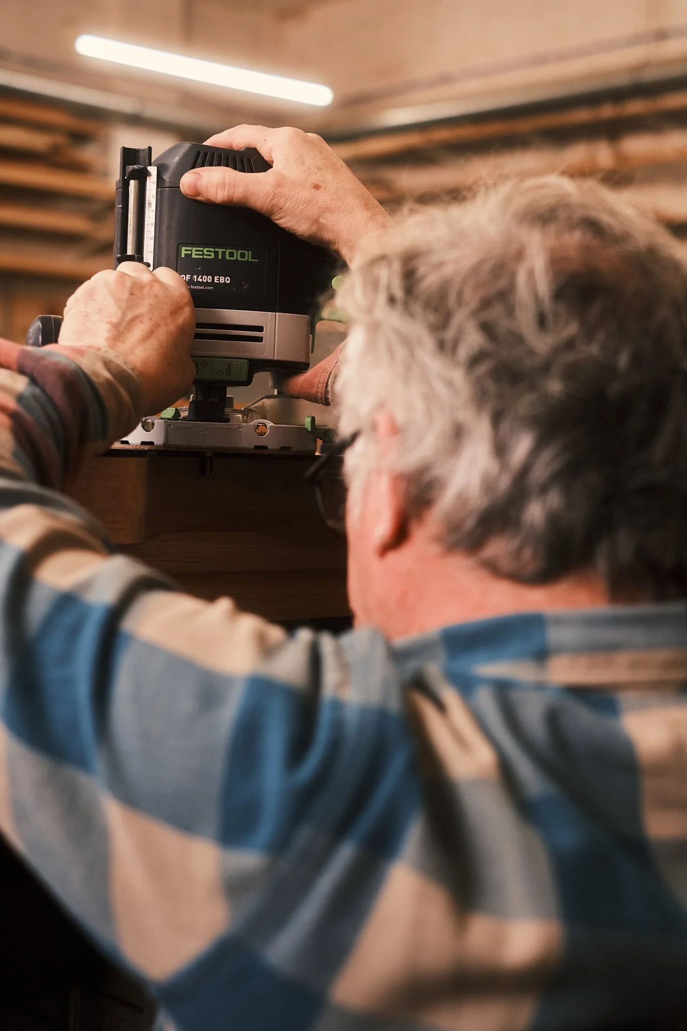 An elderly man wearing glasses and a plaid shirt is working with a Festool plunge router in a woodworking shop, focusing on his woodworking project.