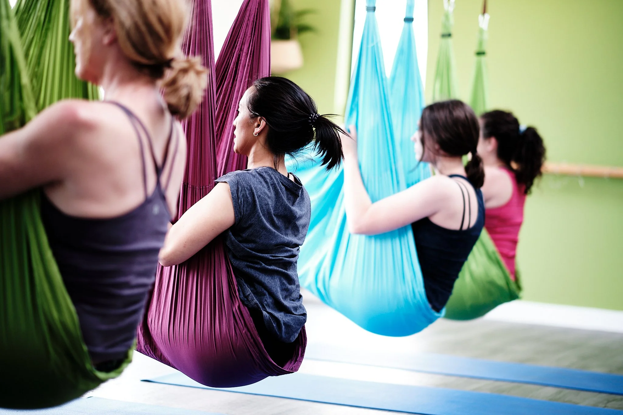 Four women practicing aerial yoga in colorful hammocks in a yoga studio.
