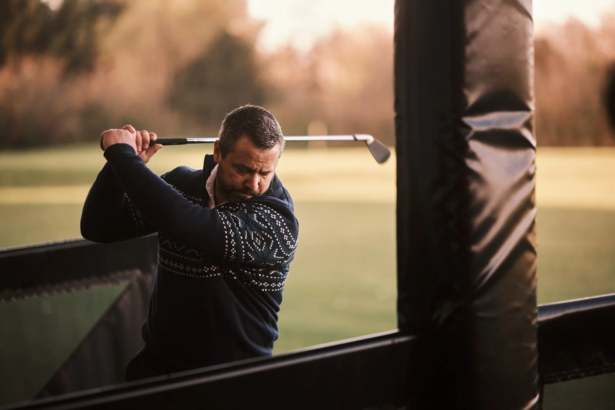 A man with short hair and a beard swinging a golf club at a golf course during sunset.