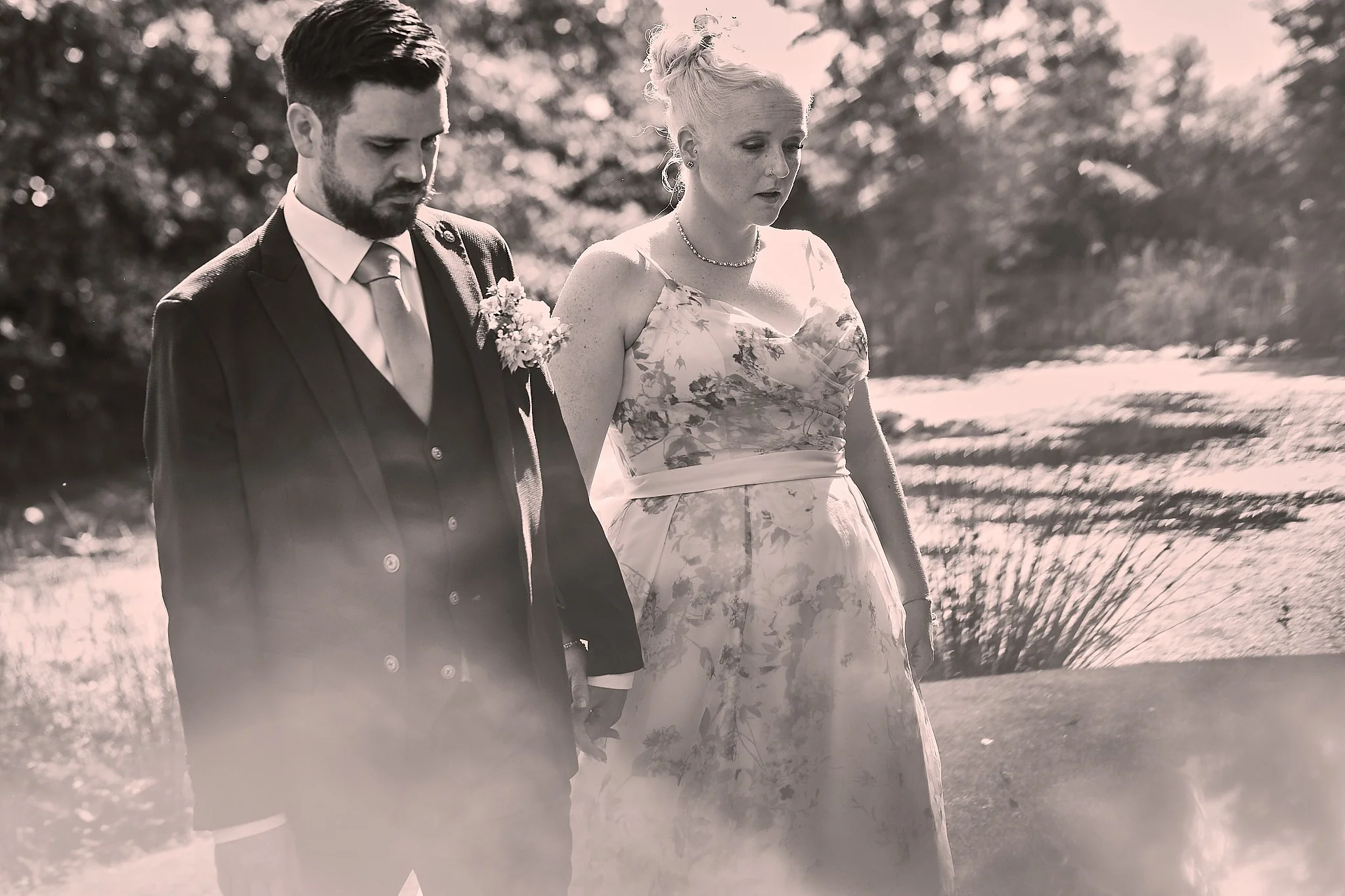 A black-and-white photo of a man in a suit and a woman in a floral dress walking outdoors, holding hands. The man has a beard and is looking down slightly, while the woman has light hair styled up and a serious expression, looking ahead. They are sur