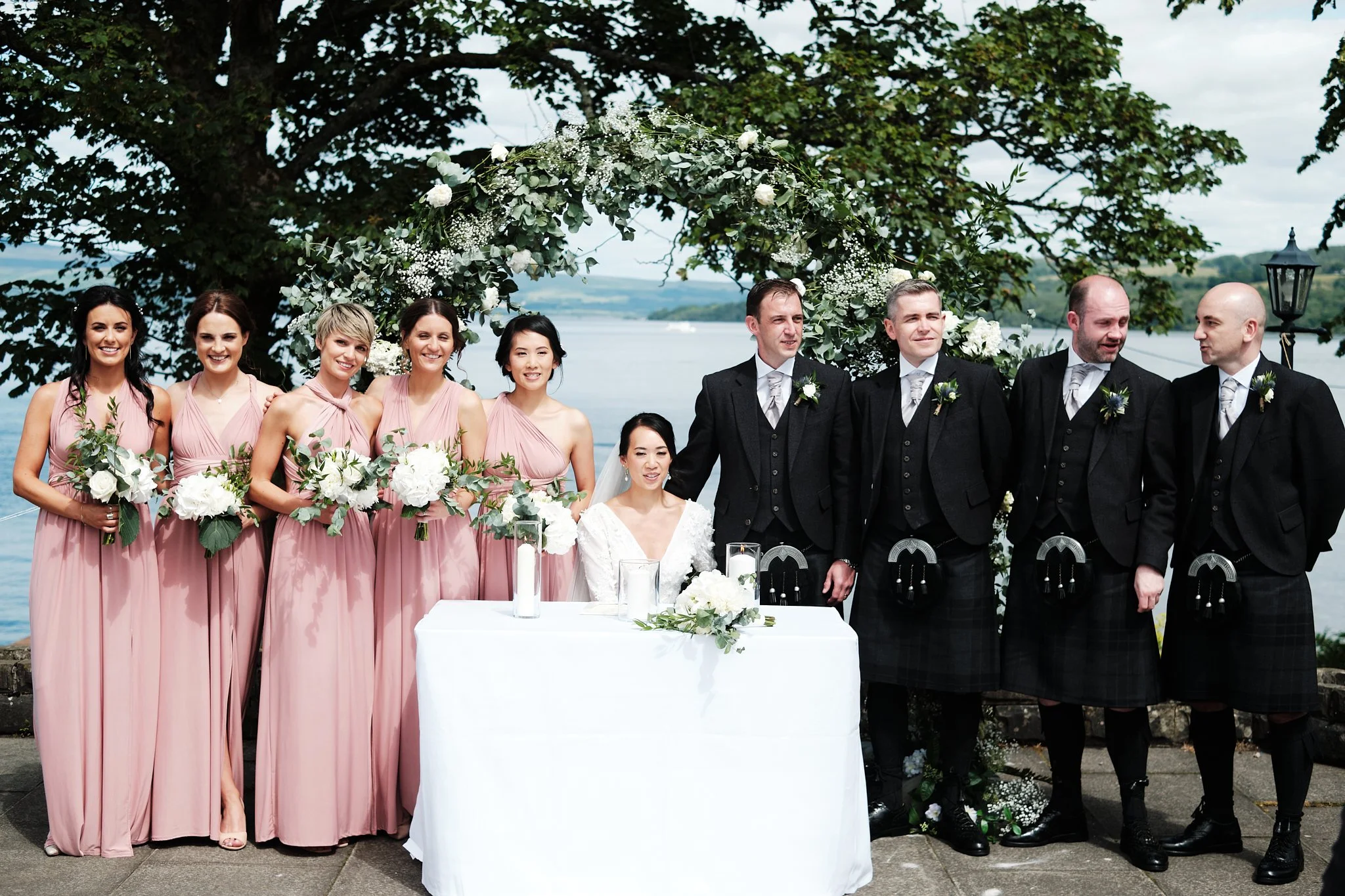 A group of wedding guests standing outdoors near a lake with a decorative floral arch overhead. The women are in pink dresses and the men in black suits, all smiling.