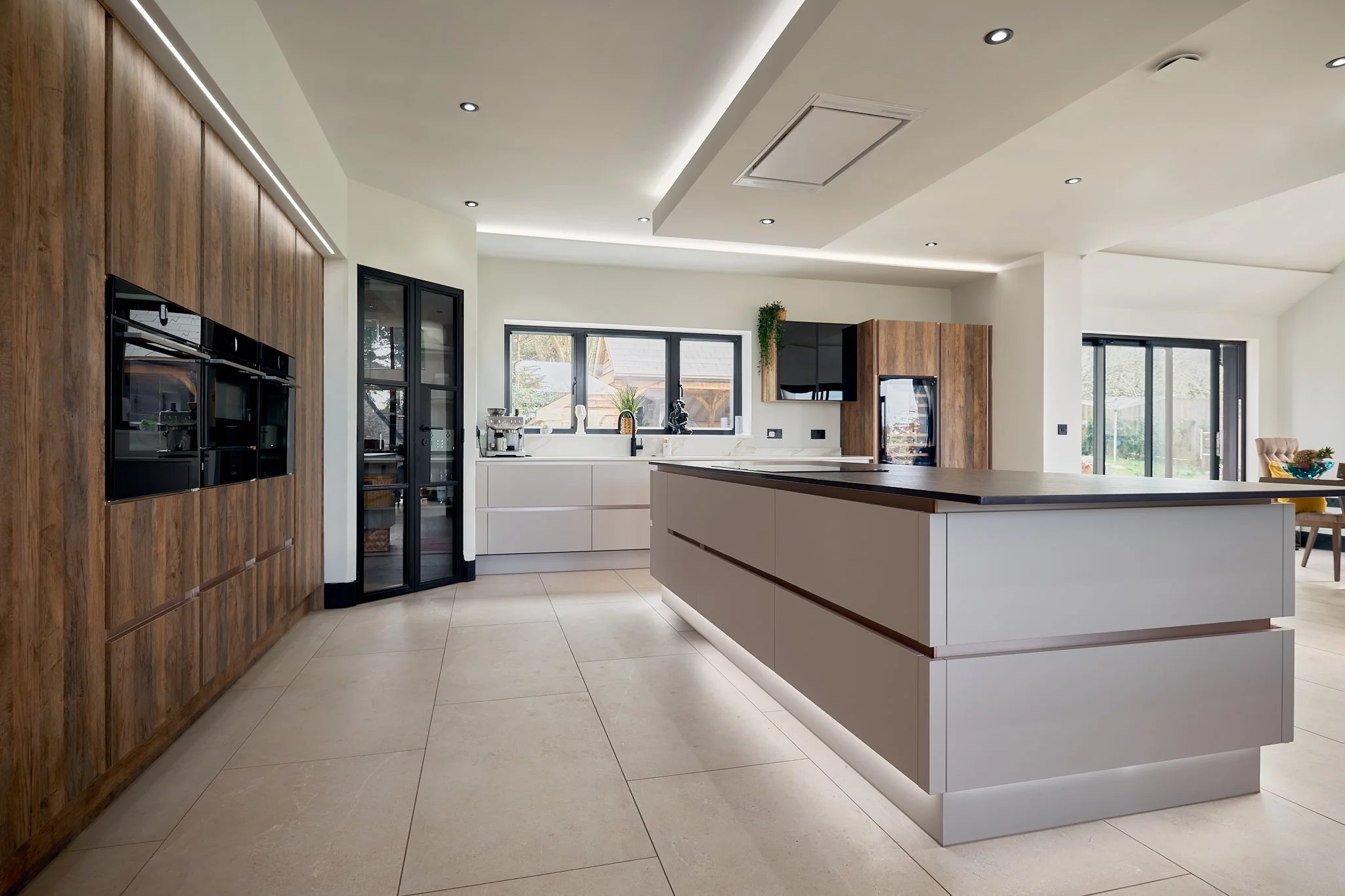 Modern kitchen with white cabinets, a large island with a dark countertop, wooden accents on the walls, and large windows letting in natural light.