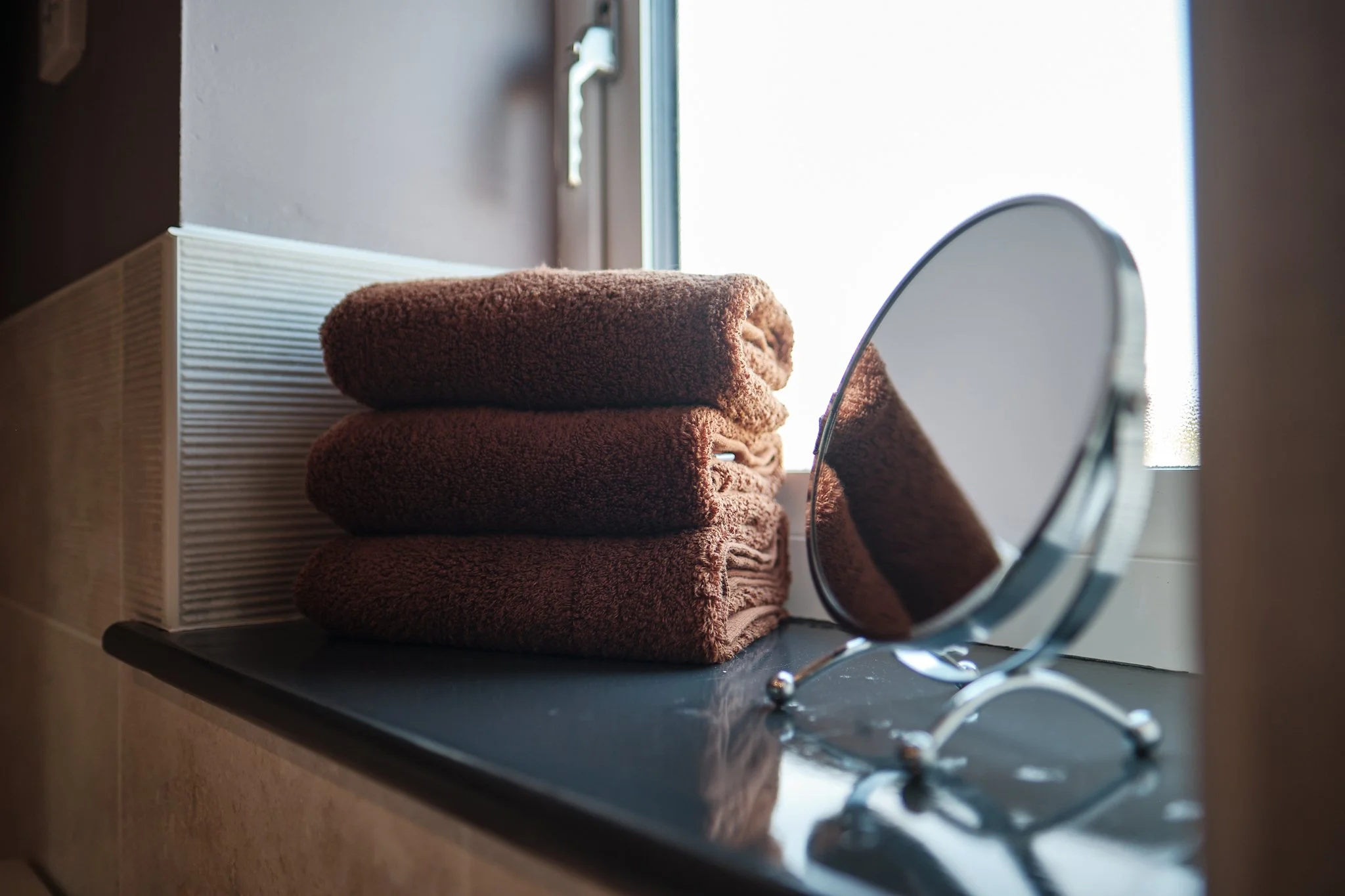 Three folded brown towels stacked on a black surface next to a round mirror and a white tissue box on a windowsill.