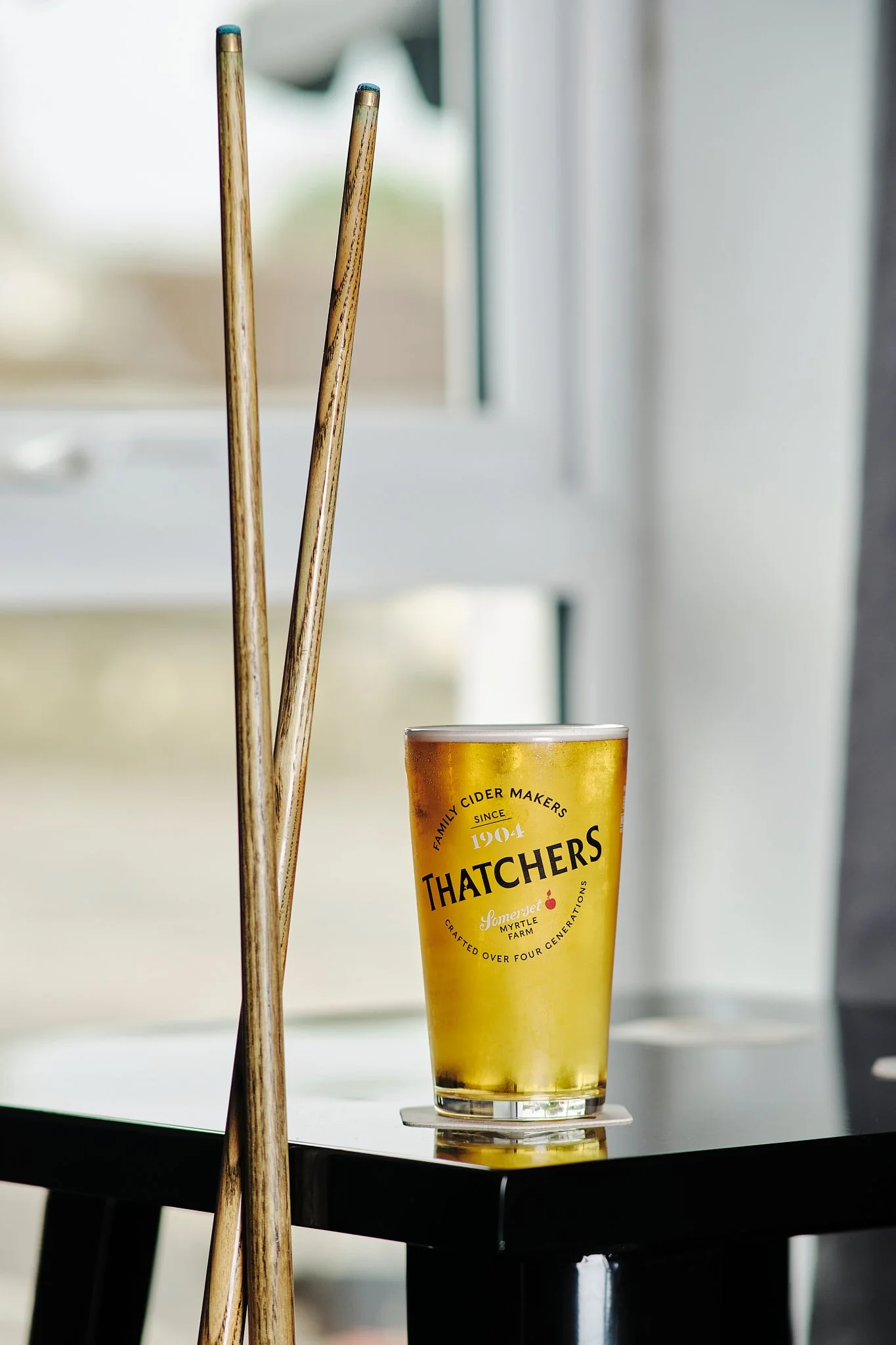 A glass of Thatchers cider on a table with a coaster, with two wooden chopsticks leaning against each other nearby, near a window with blurred background.