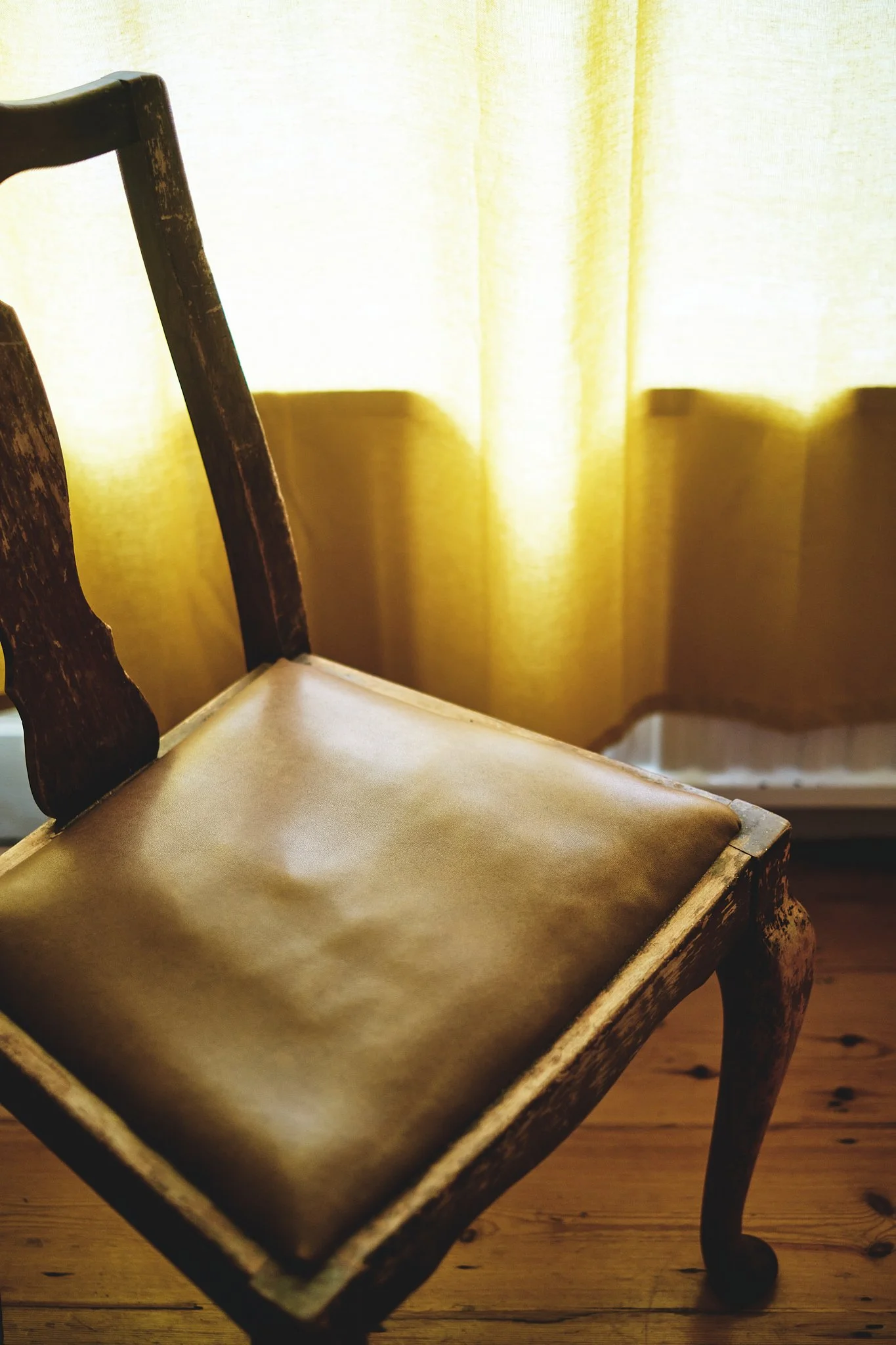 An antique wooden chair with a padded leather seat, placed in front of a yellow curtain with light shining through.