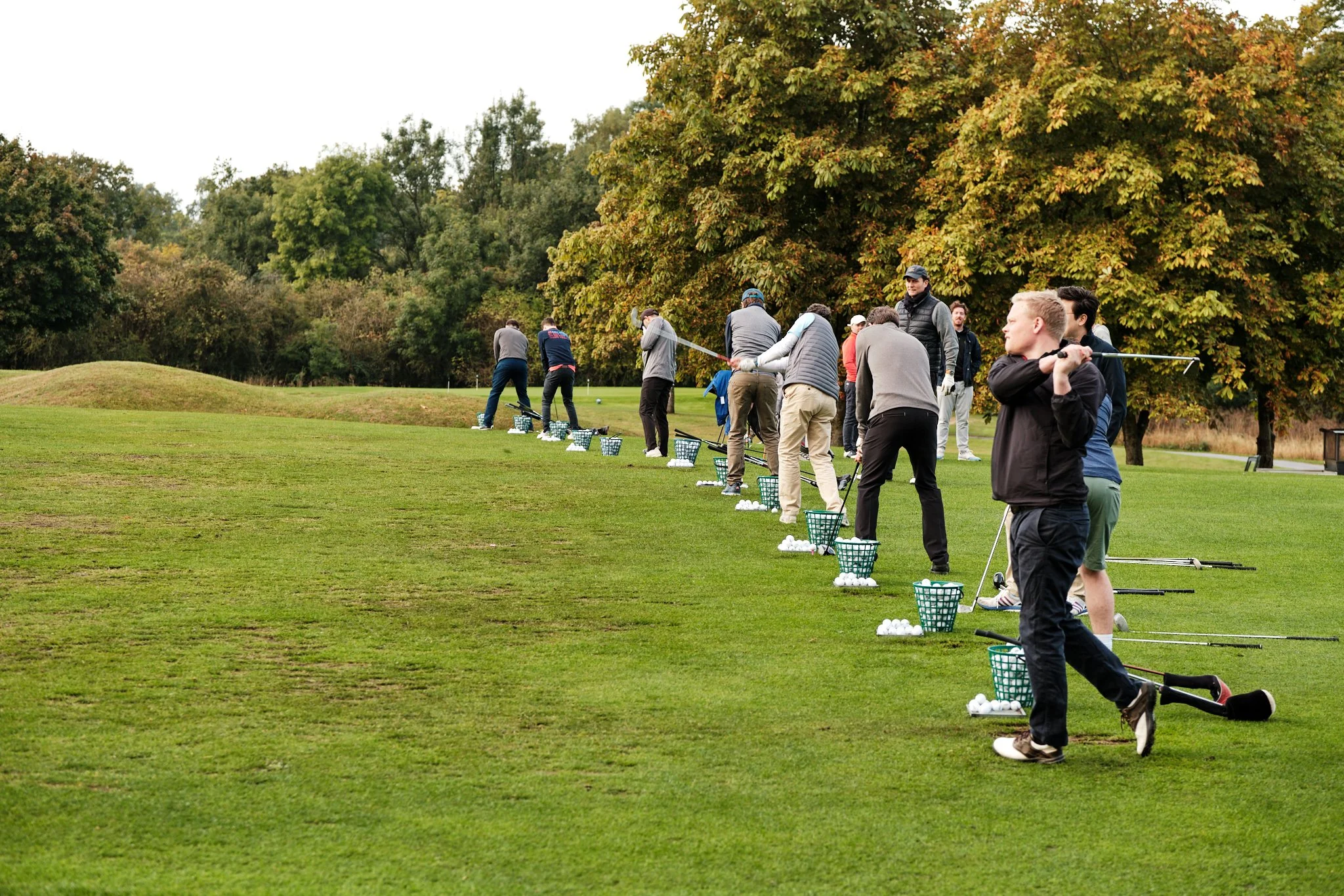 Group of people practicing golf on a practice range with trees in the background.