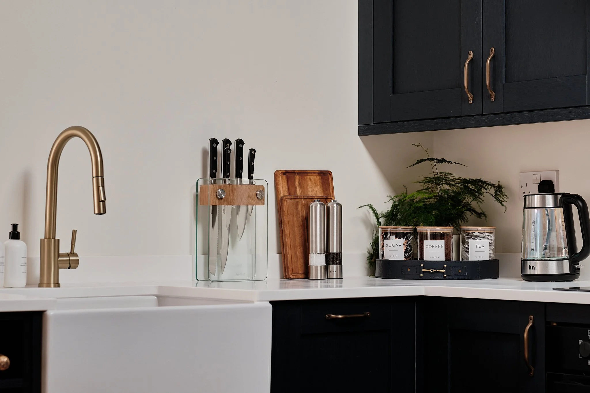 Kitchen countertop with black upper cabinets and brass handles, gold faucet, knives in a glass block, wooden cutting boards, salt and pepper shakers, plants, containers labeled sugar, coffee, tea, and a kettle.