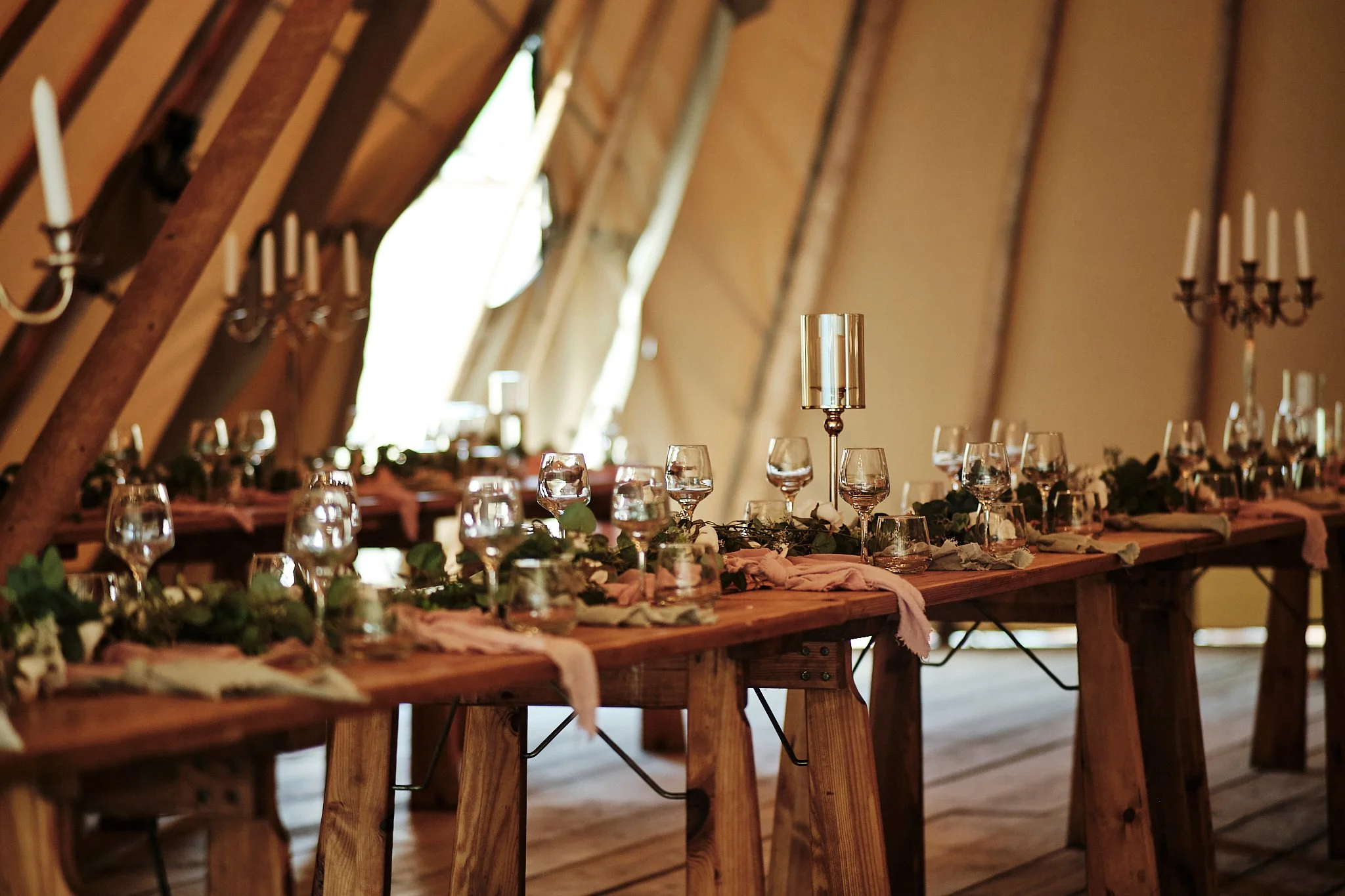 A long wooden table set for a wedding reception with glasses, candles, and floral decorations inside a rustic barn.