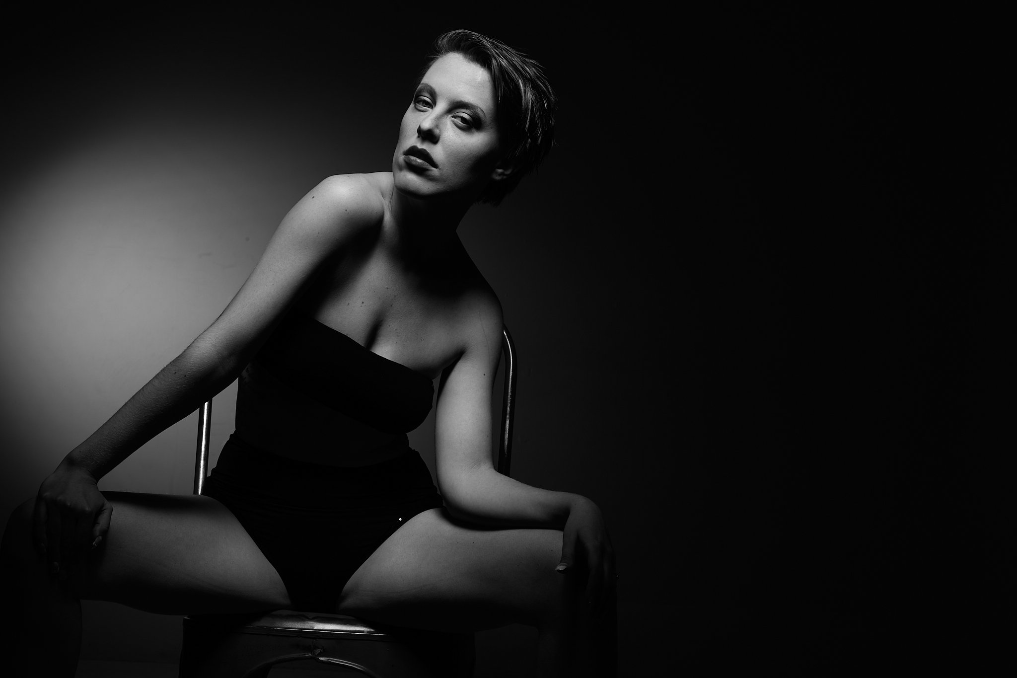 A black and white photograph of a woman sitting on a chair, wearing a strapless dress, with short hair, looking at the camera with a serious expression. The lighting highlights her face and upper body, creating a dramatic effect.