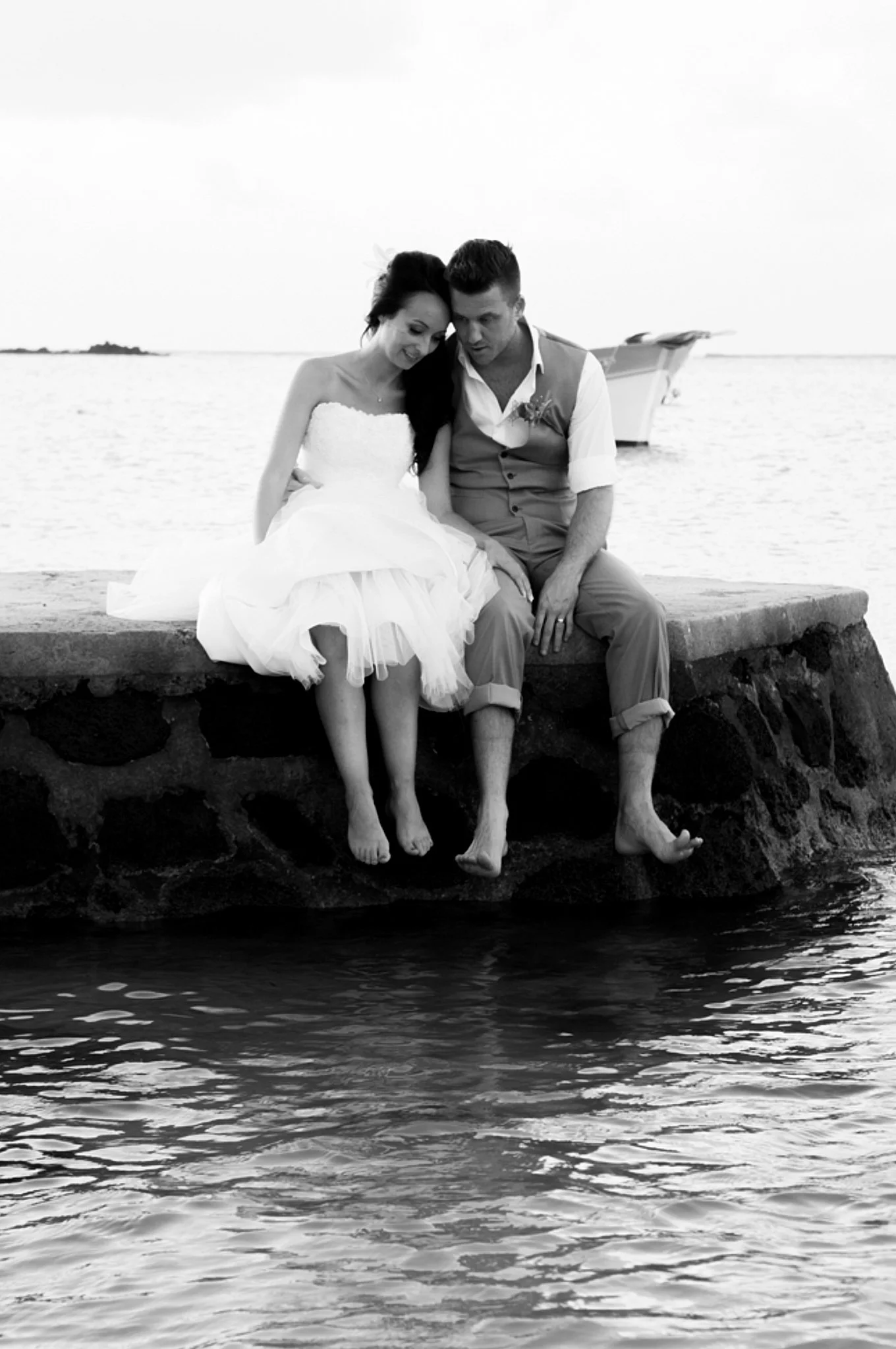 A black and white photo of a couple sitting on a stone wall by the water, with a boat visible in the background. The woman is wearing a strapless dress, and the man is dressed in a short-sleeved shirt and pants. They are sitting close, with their hea