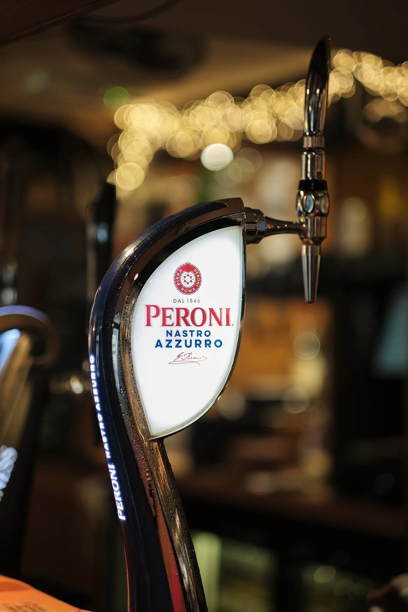 Close-up of a Peroni beer tap with the Peroni logo and the words 'Nastro Azzurro' in a bar setting with bokeh lights in the background.