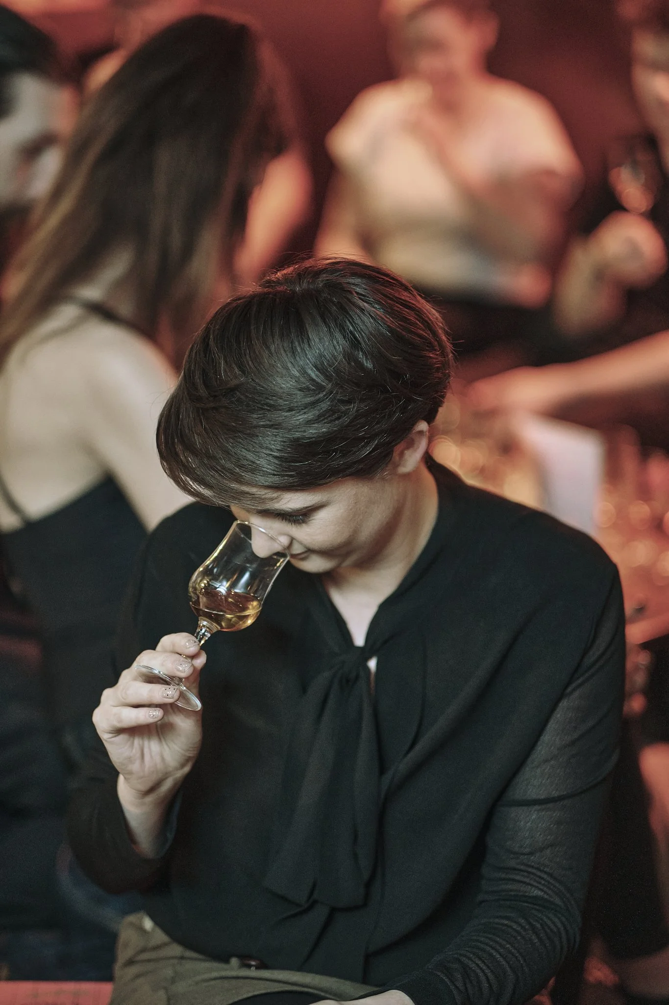 A woman with short dark hair and a black shirt is holding a wine glass, smelling the drink in a dimly lit, lively social setting with other people in the background.