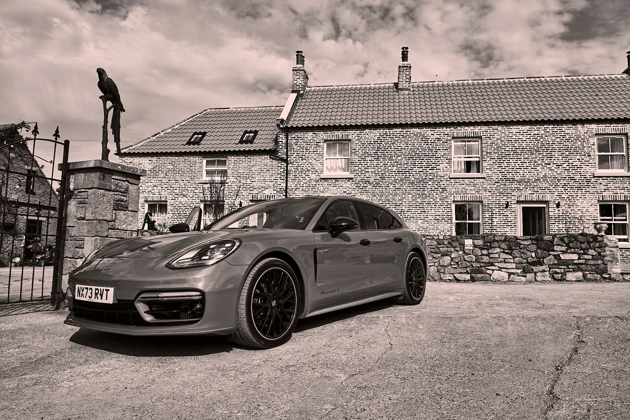 A modern, silver sports car parked in front of a brick building with brick chimneys and a tiled roof. The sky is cloudy.