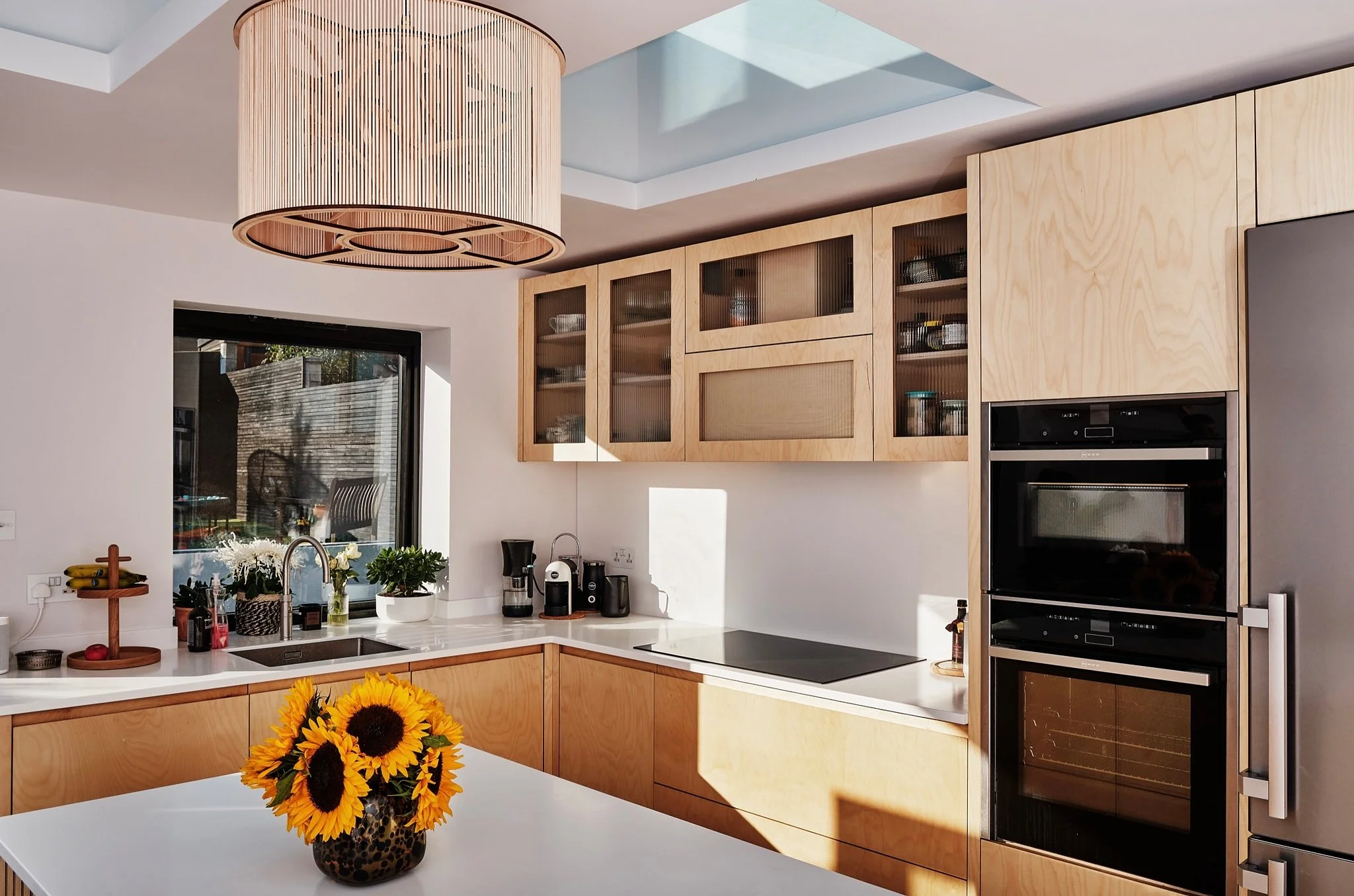 Modern kitchen with light wood cabinets, a window, a white countertop, a stovetop, two built-in ovens, and a vase of sunflowers on a white table. Sunlight streams through a skylight.