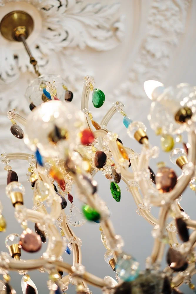 Close-up of a decorative chandelier with colorful crystals hanging from gold and clear branches, with an ornate white ceiling in the background.