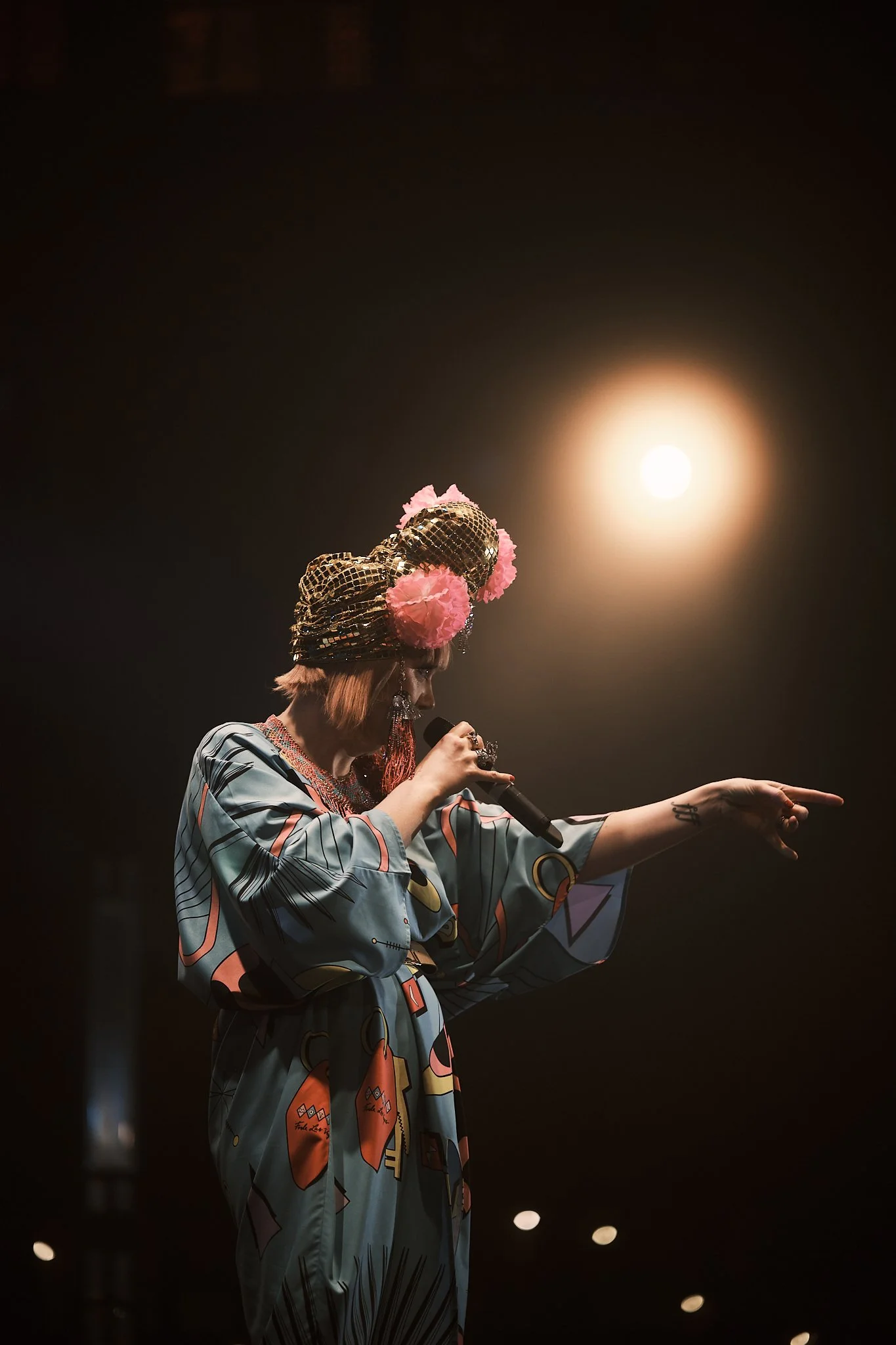 A performer with pink hair, wearing a patterned silvery head wrap and large pink floral decorations, singing into a microphone on stage, illuminated by a bright light and dressed in a colorful, abstract patterned outfit.